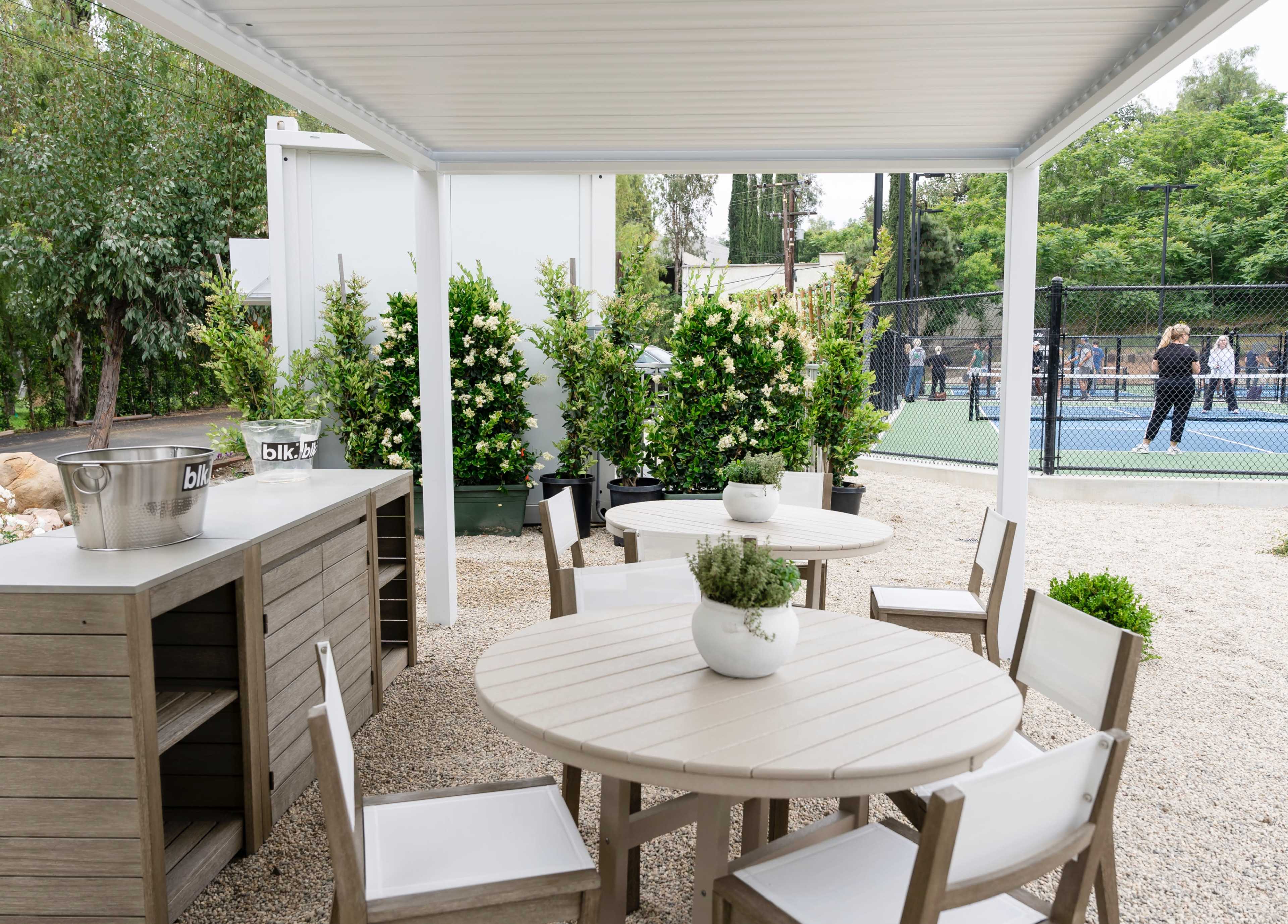 A shaded outdoor seating area features wooden tables and chairs, surrounded by manicured plants, with a tennis court visible in the background.