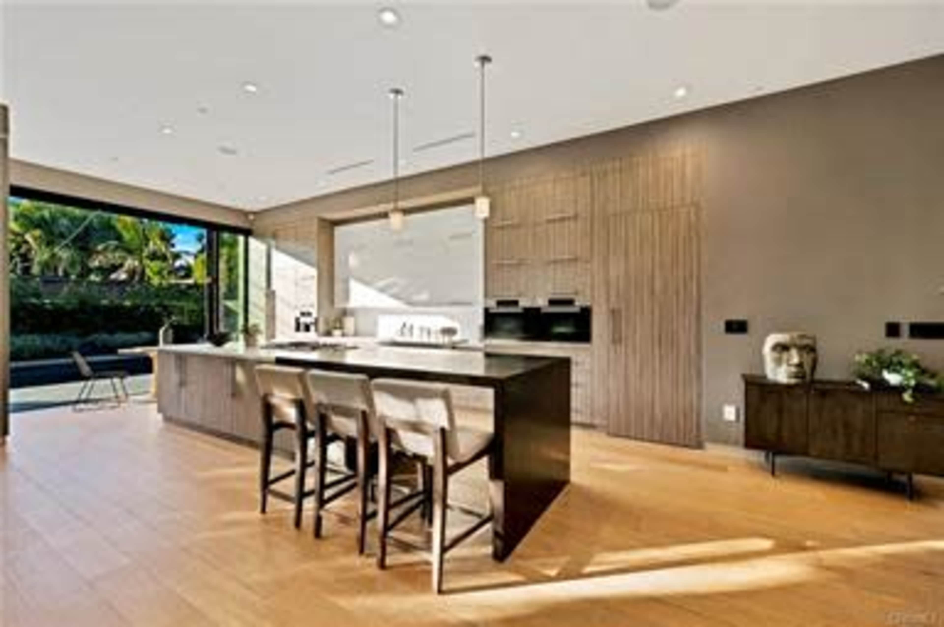 A modern kitchen with a large island, three bar stools, and floor-to-ceiling windows that open to a patio.