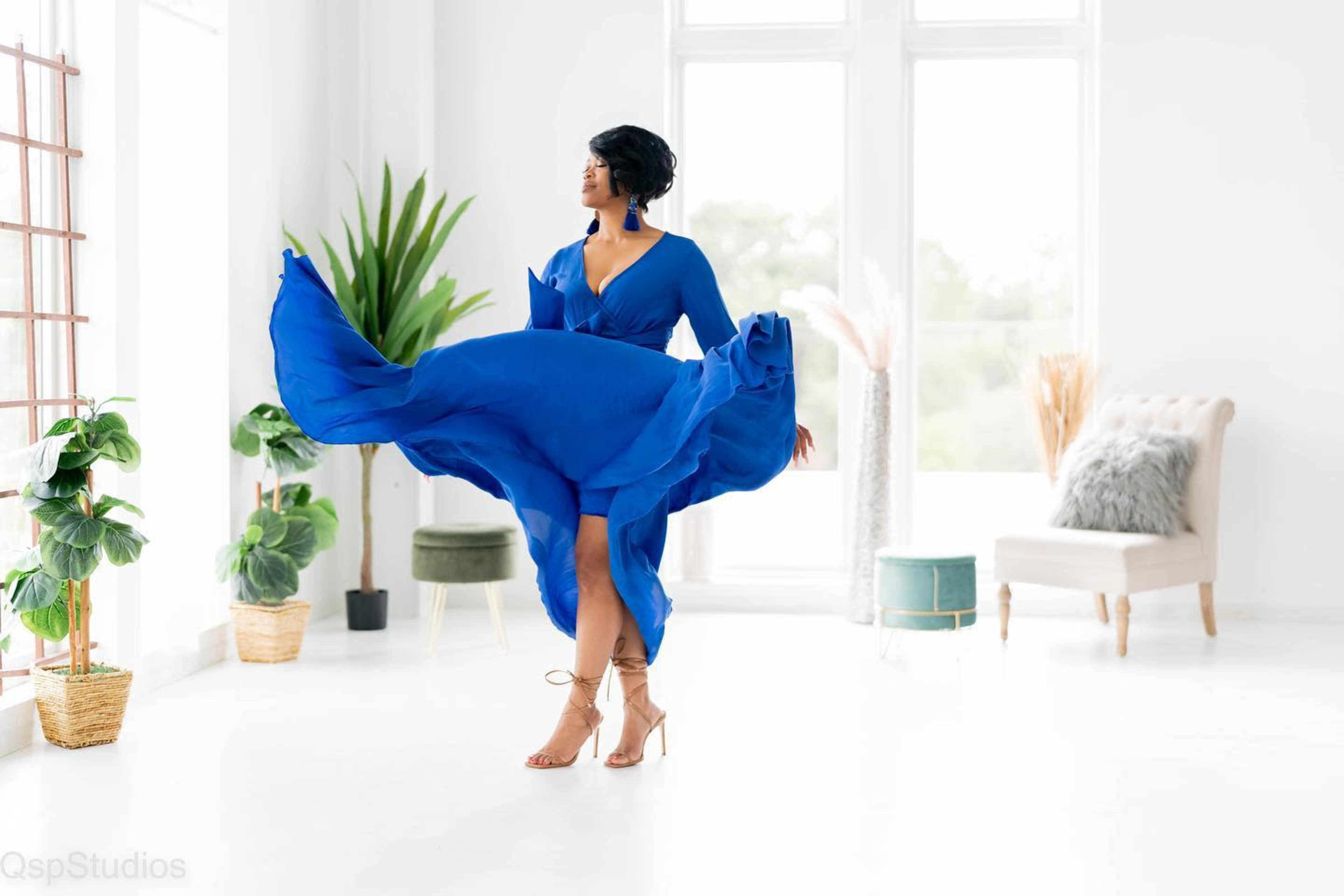 A woman in a blue dress twirls in a bright, airy room with large windows and potted plants.