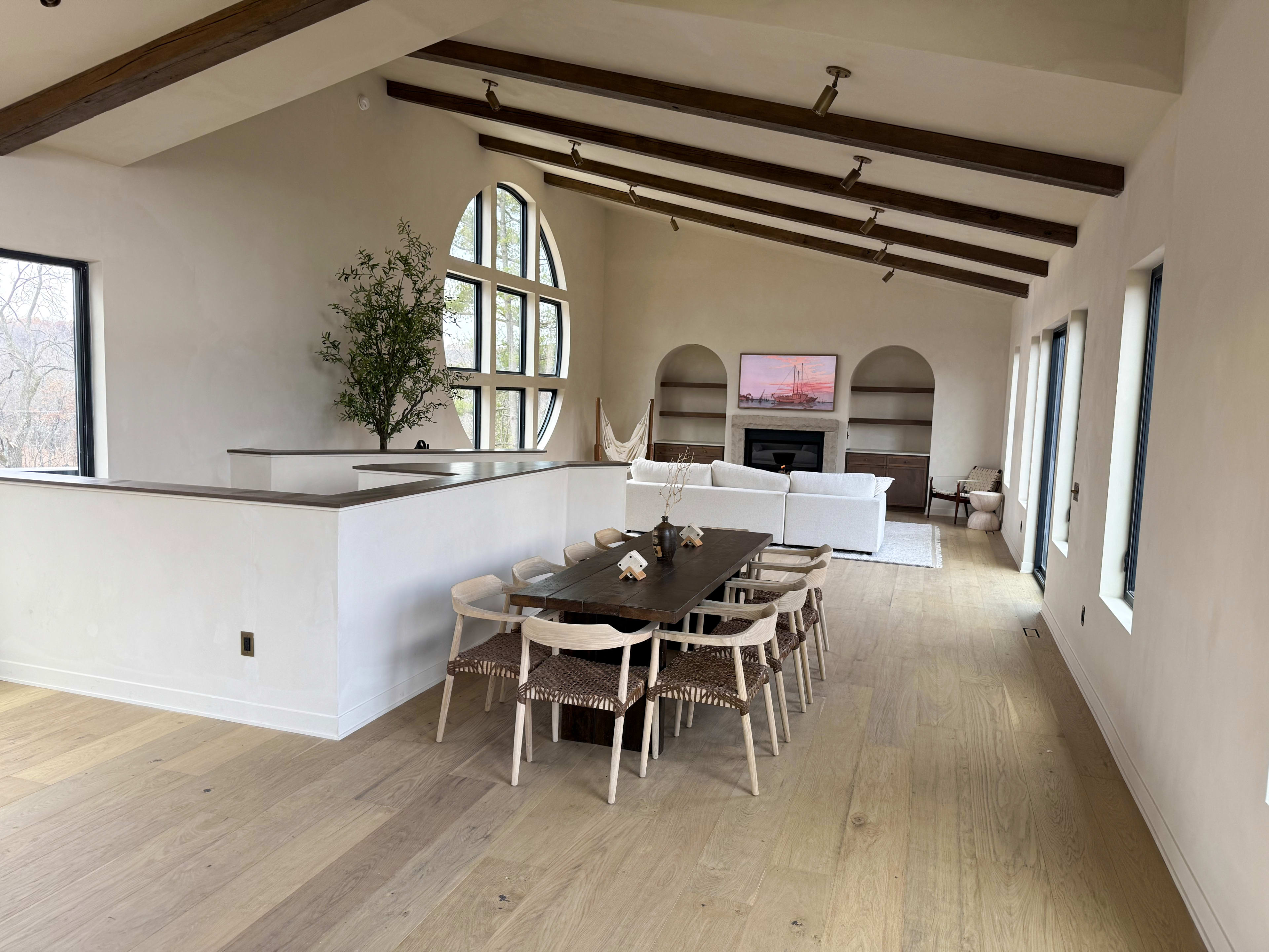A spacious interior features a dining area with a long table surrounded by chairs, and a living room area with a white sofa and a television mounted on the wall.
