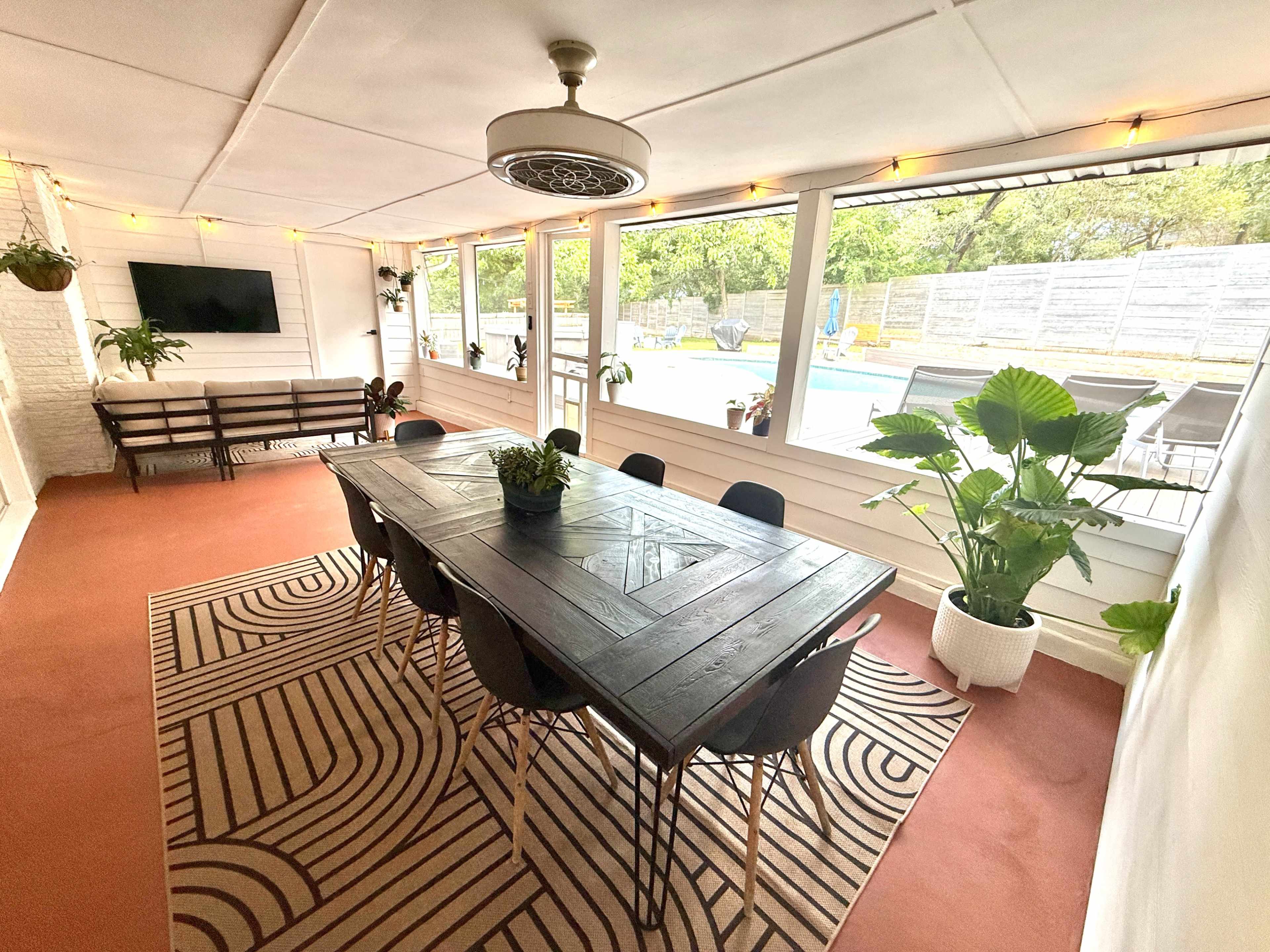 The image shows a sunlit dining area with a long rectangular table surrounded by black chairs, large windows providing a view of an outdoor pool, and houseplants arranged around the space.
