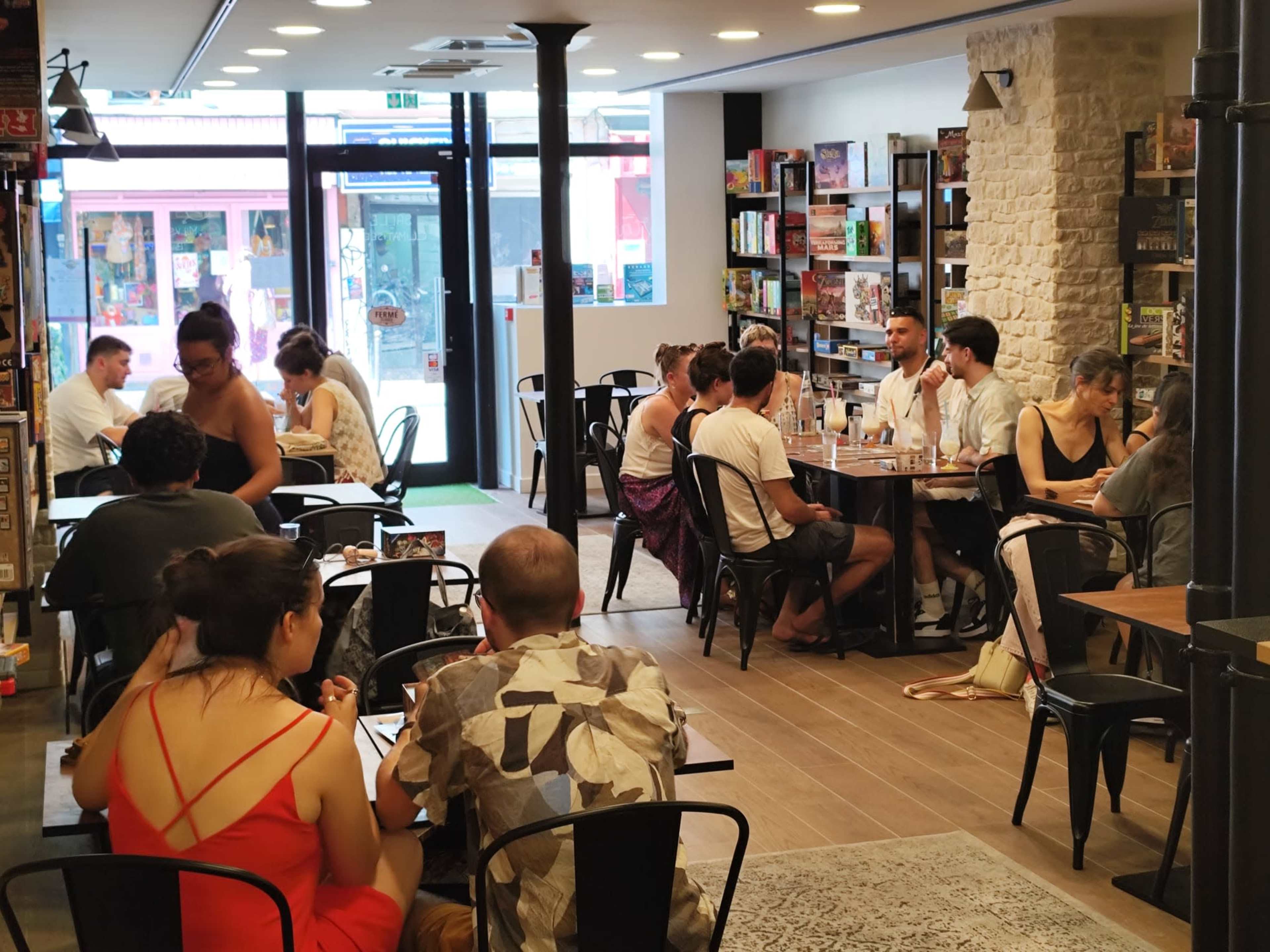 The image shows a busy board game café with groups of people sitting around tables engaged in games or conversation.
