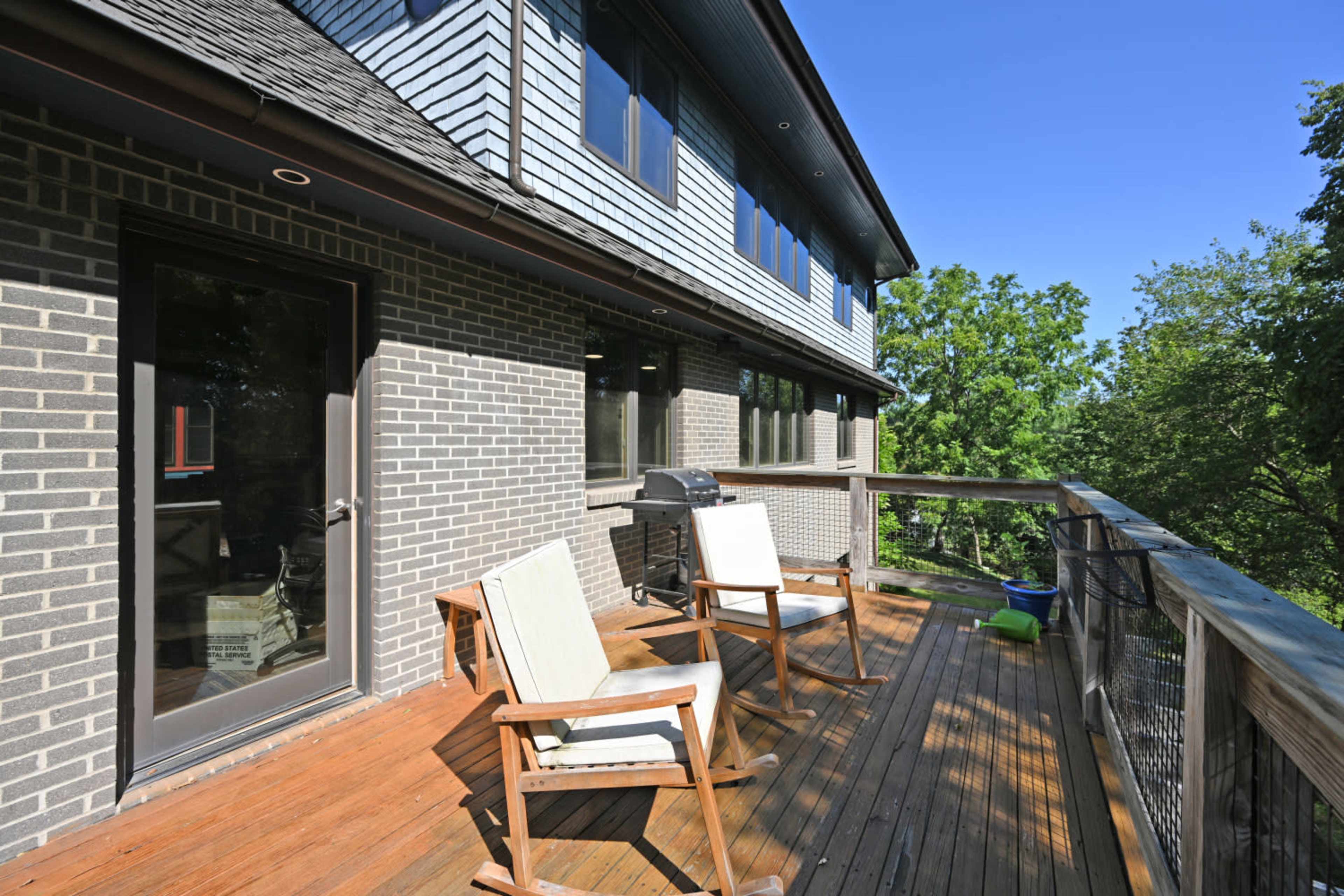 The image shows a wooden deck with two chairs and a barbecue grill, attached to a brick house surrounded by greenery.
