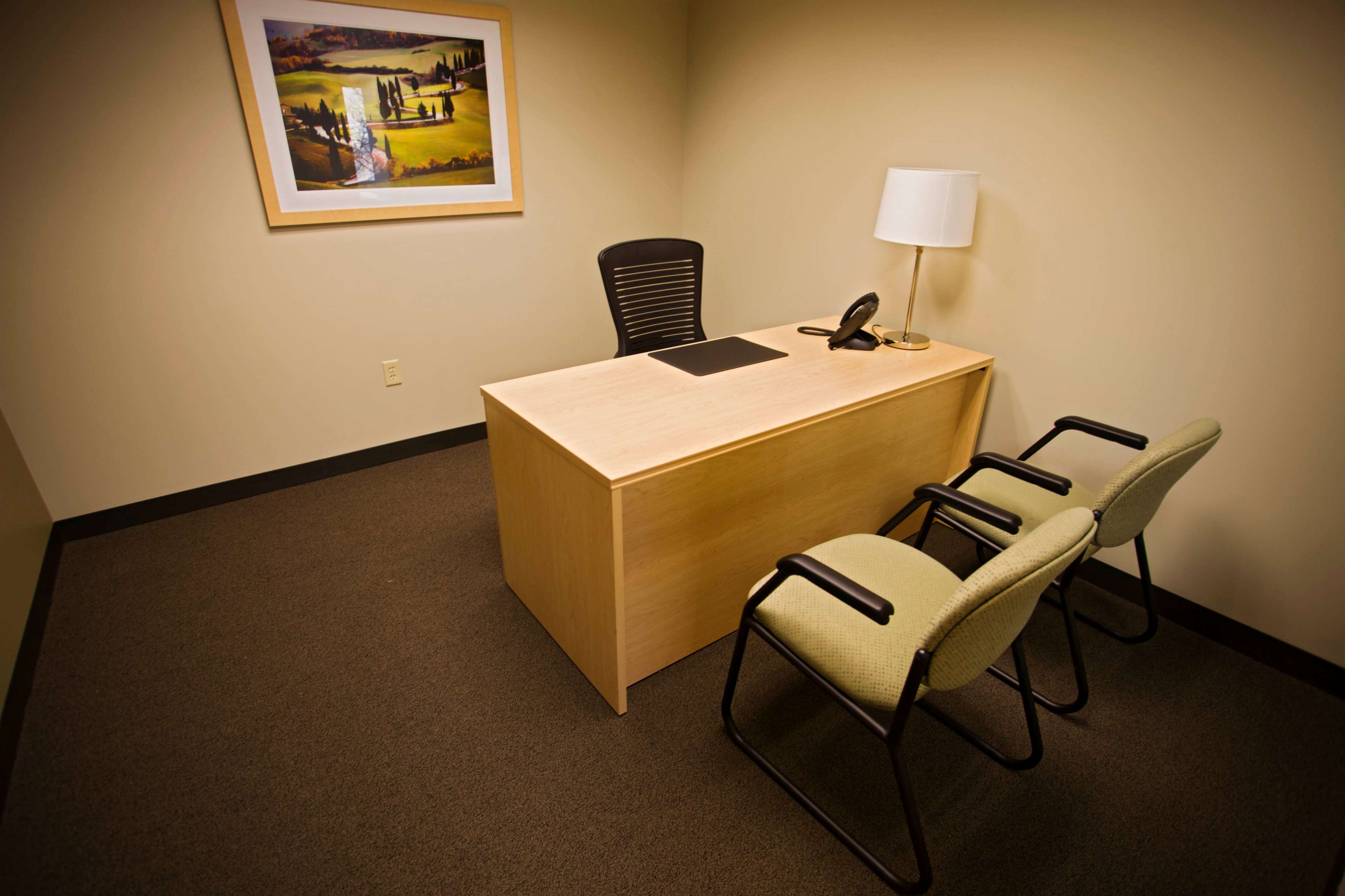 A simple office space features a desk with a phone, a lamp, and two chairs, alongside a framed landscape painting on the wall.