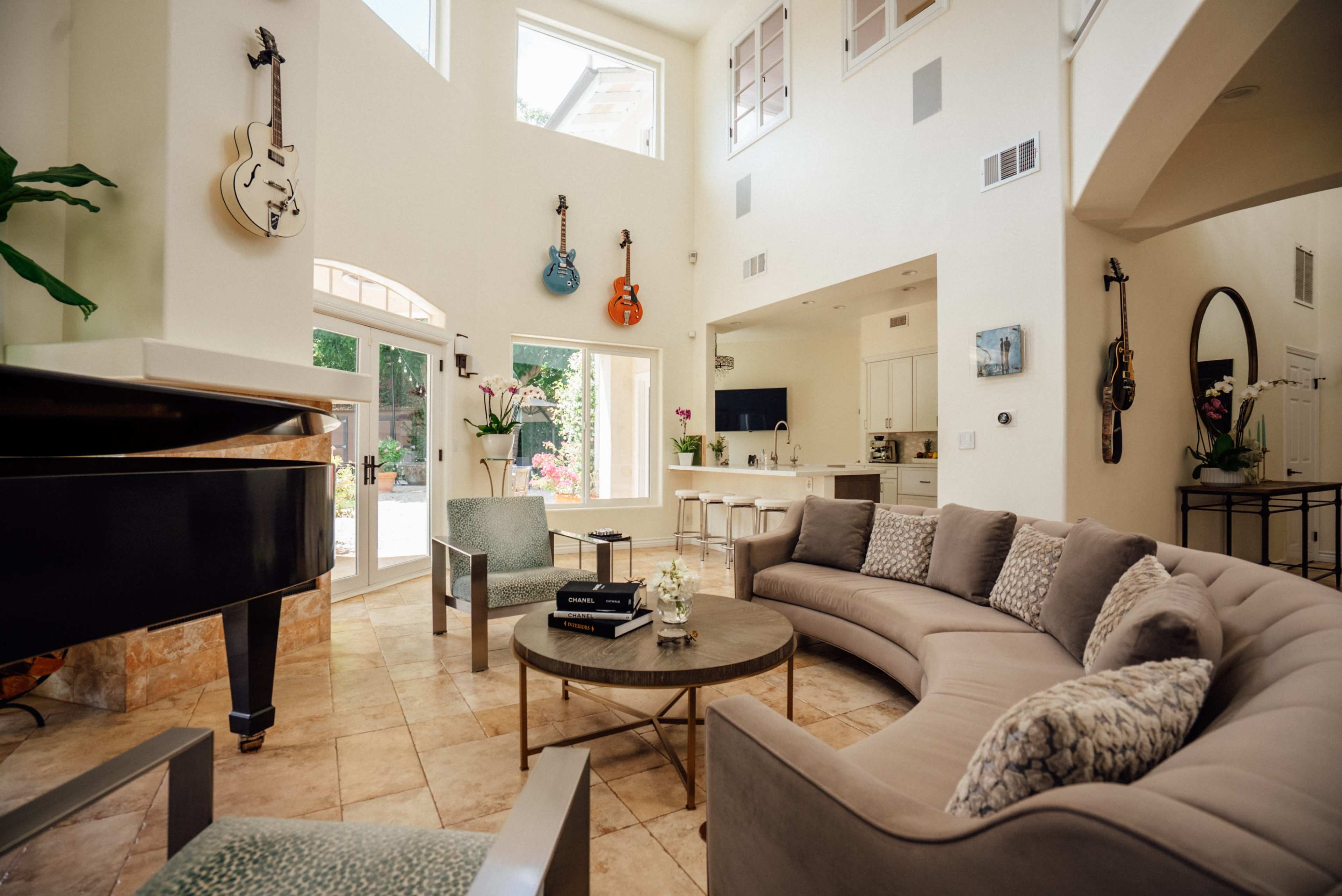 A spacious living room features a curved sectional sofa, a grand piano, and guitars hanging on the walls, with large windows allowing natural light to fill the space.