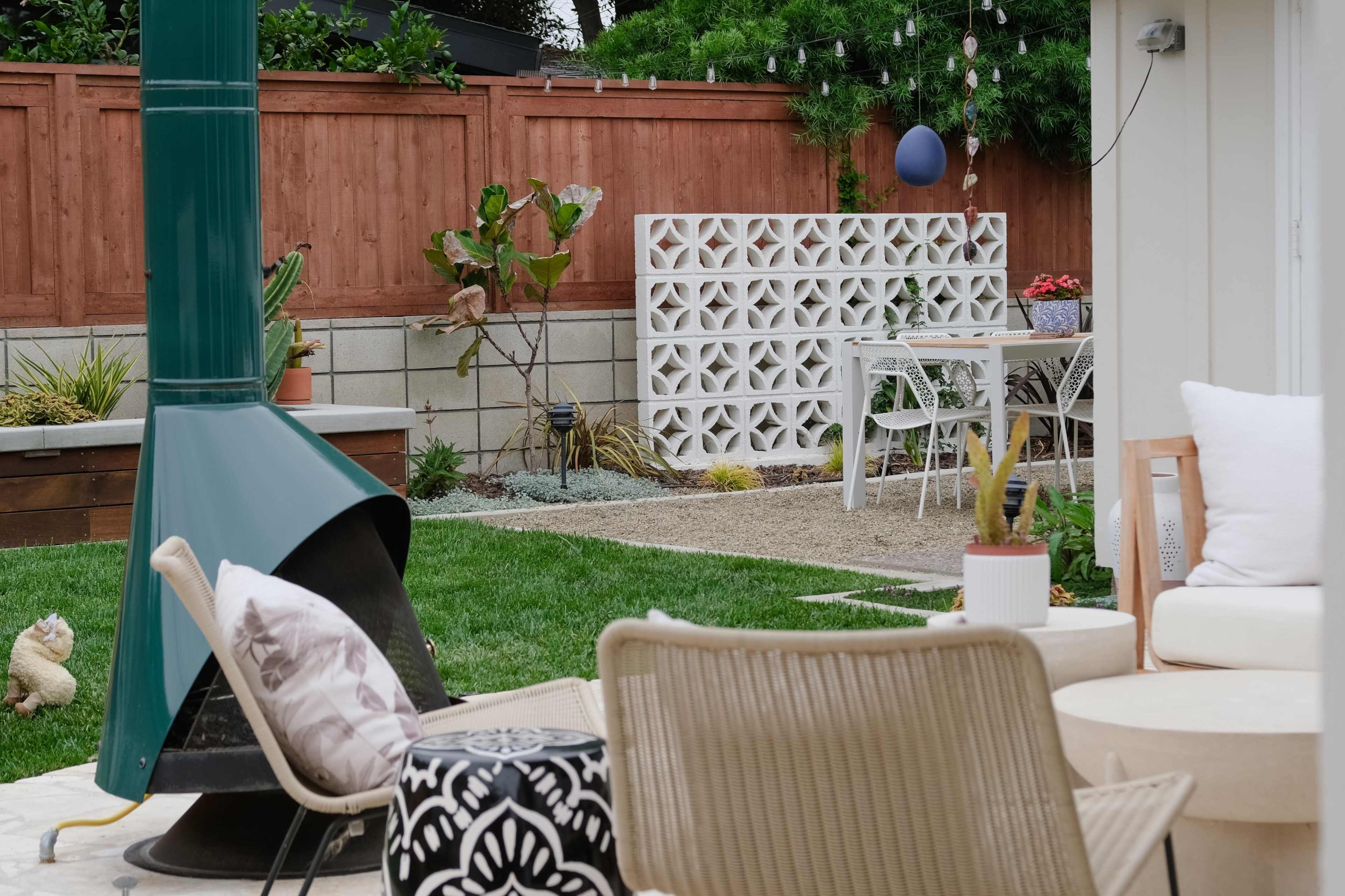The image shows a landscaped backyard featuring a seating area with a fire pit, decorative plants, and a dining table behind a decorative concrete screen.