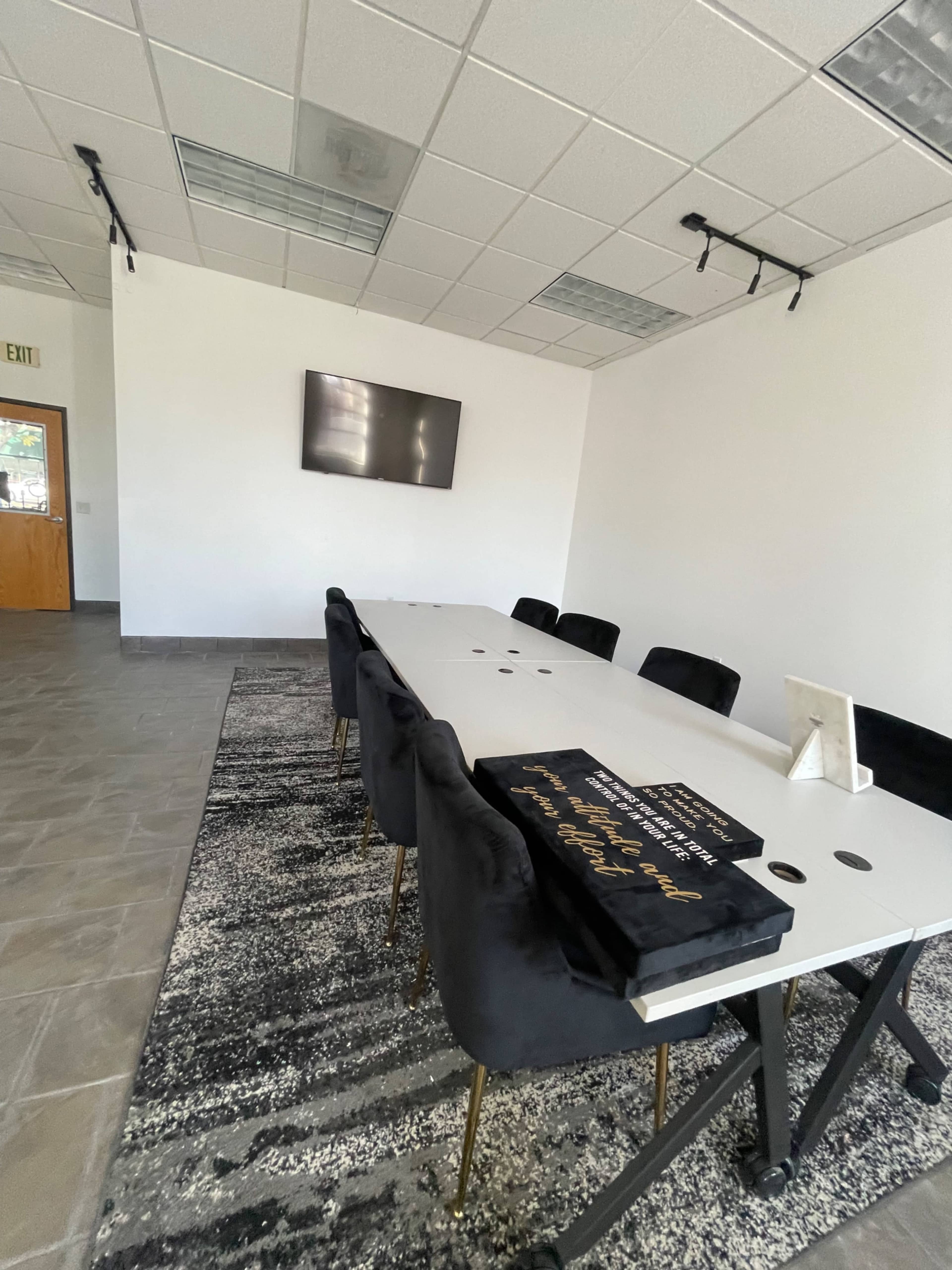 A modern conference room features a long table surrounded by black chairs, with a large screen on the wall and a rug on the floor.