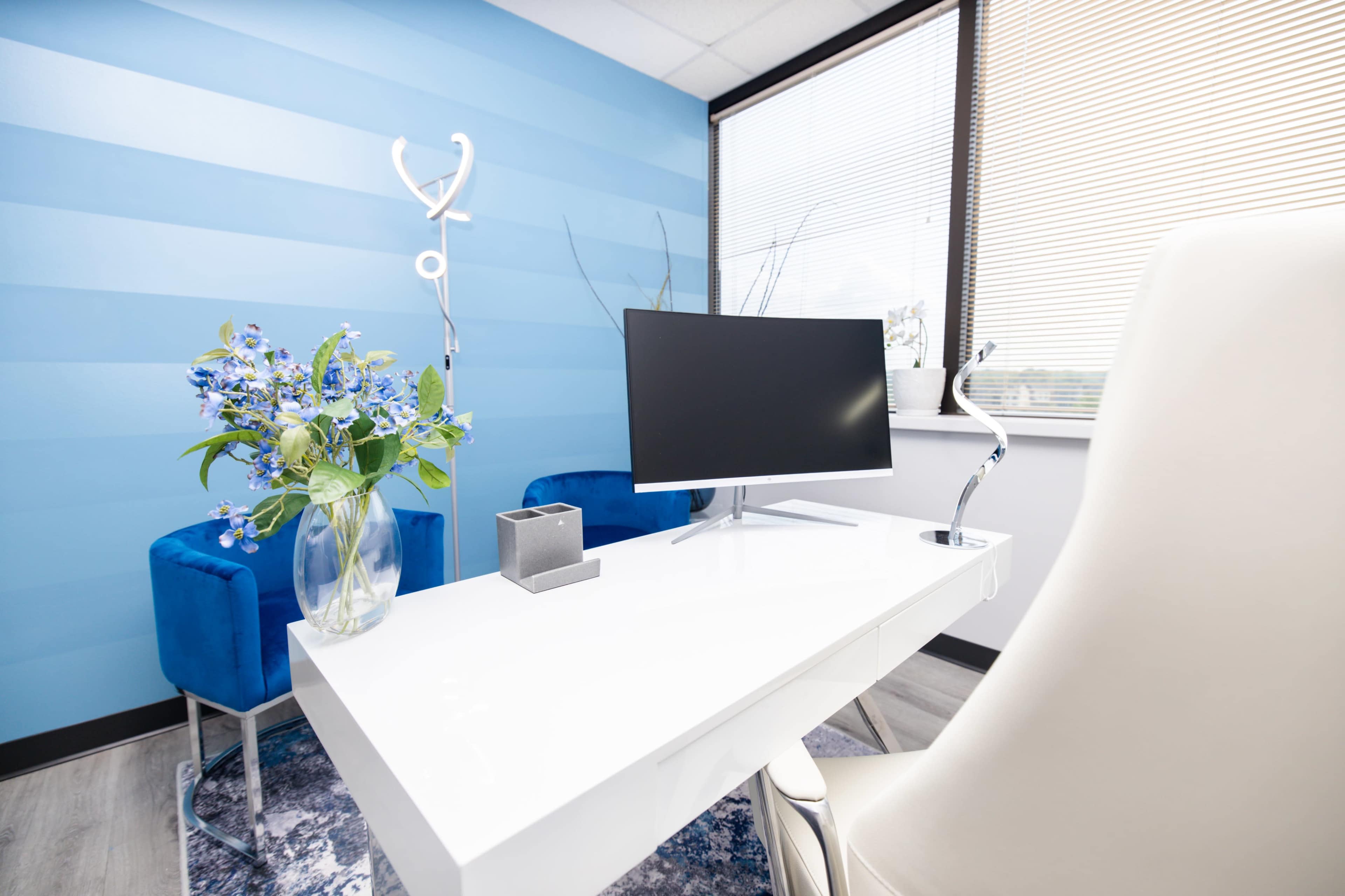 A modern office space features a white desk with a computer monitor, blue chairs, and a vase of blue flowers against a striped blue wall.