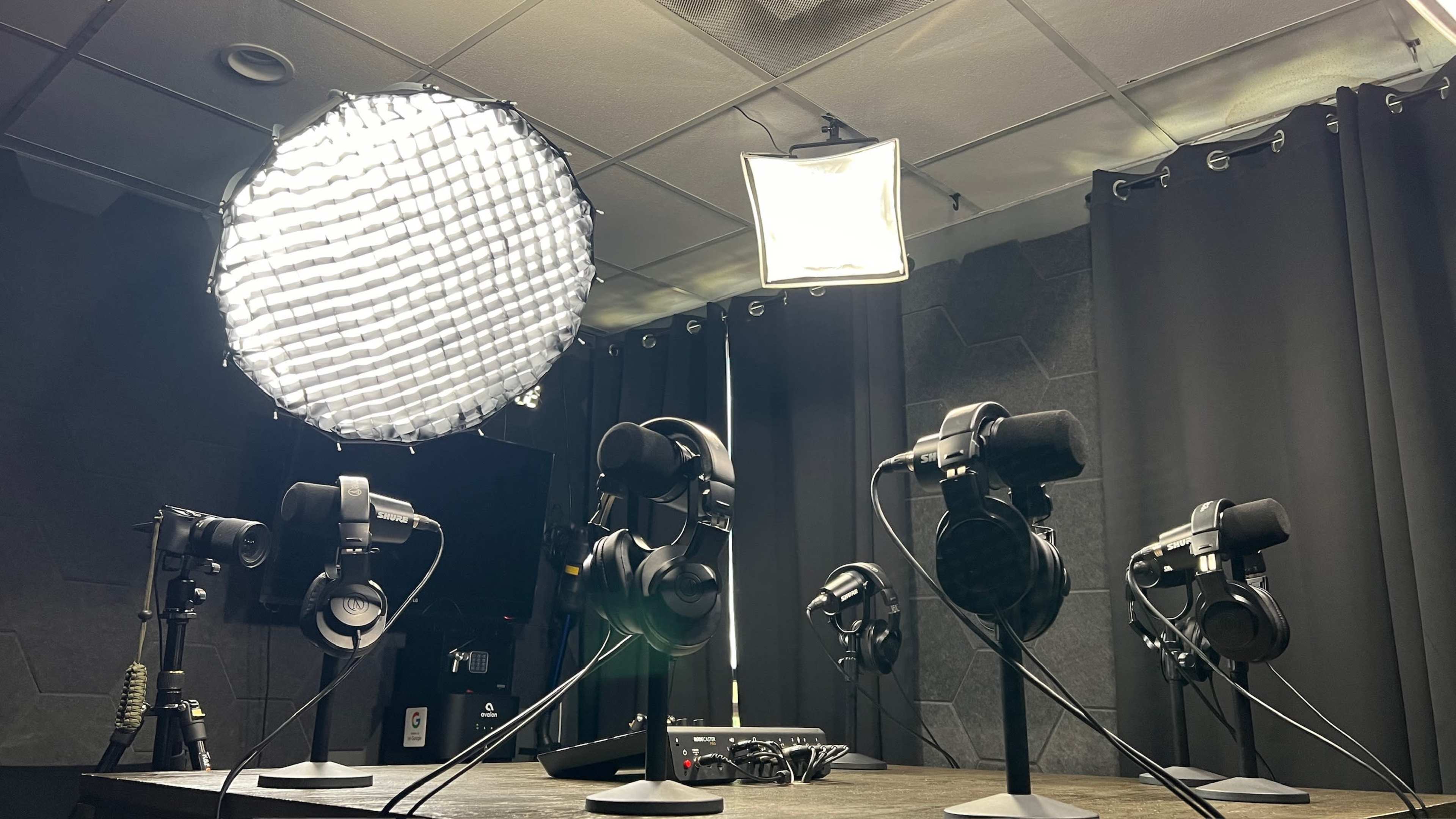 A recording studio setup features multiple microphones and headphones arranged on stands, illuminated by overhead lights against a backdrop of dark curtains.
