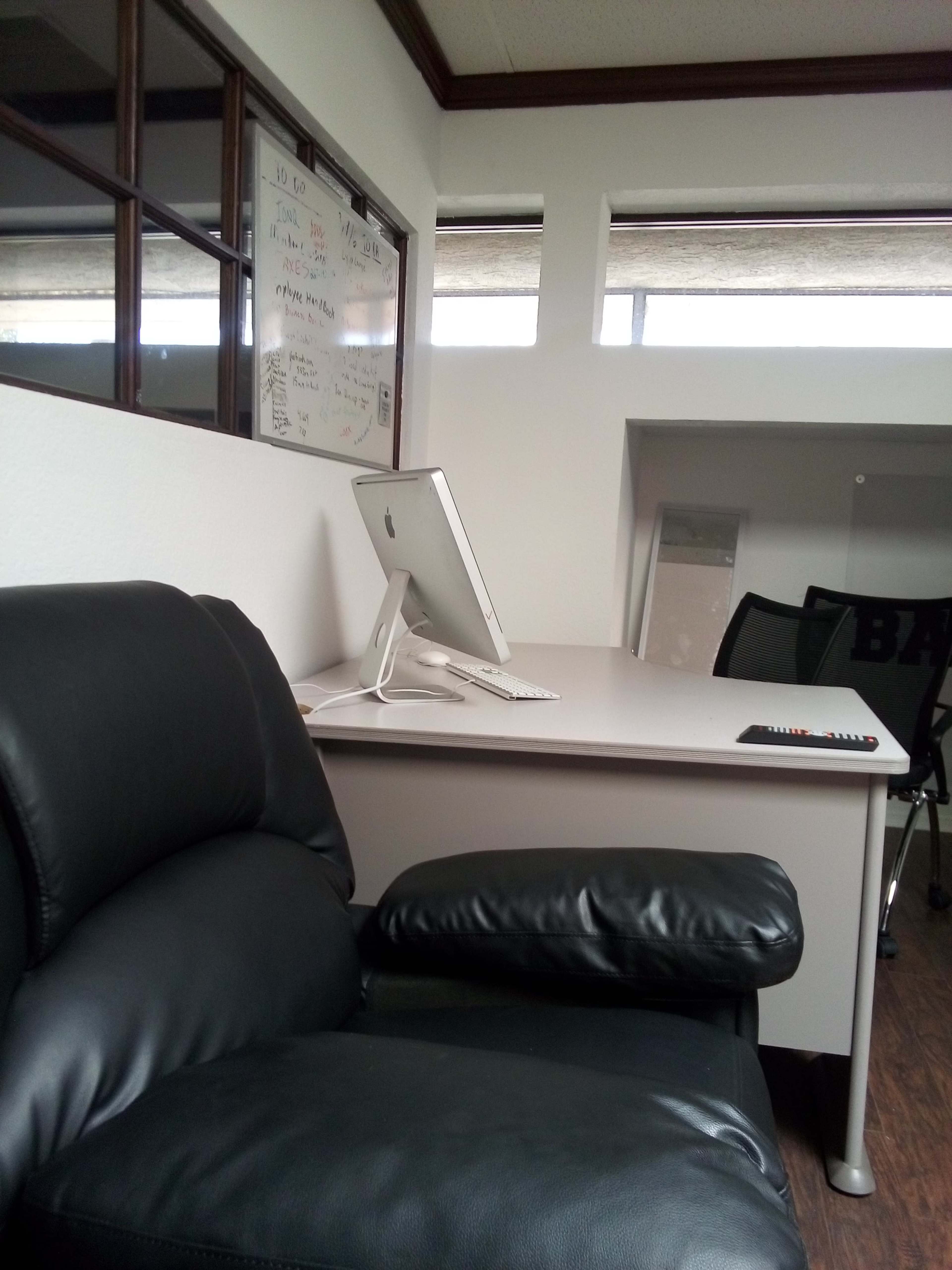 The image shows a modern office space featuring a black leather chair beside a white desk with a computer monitor and a remote control.