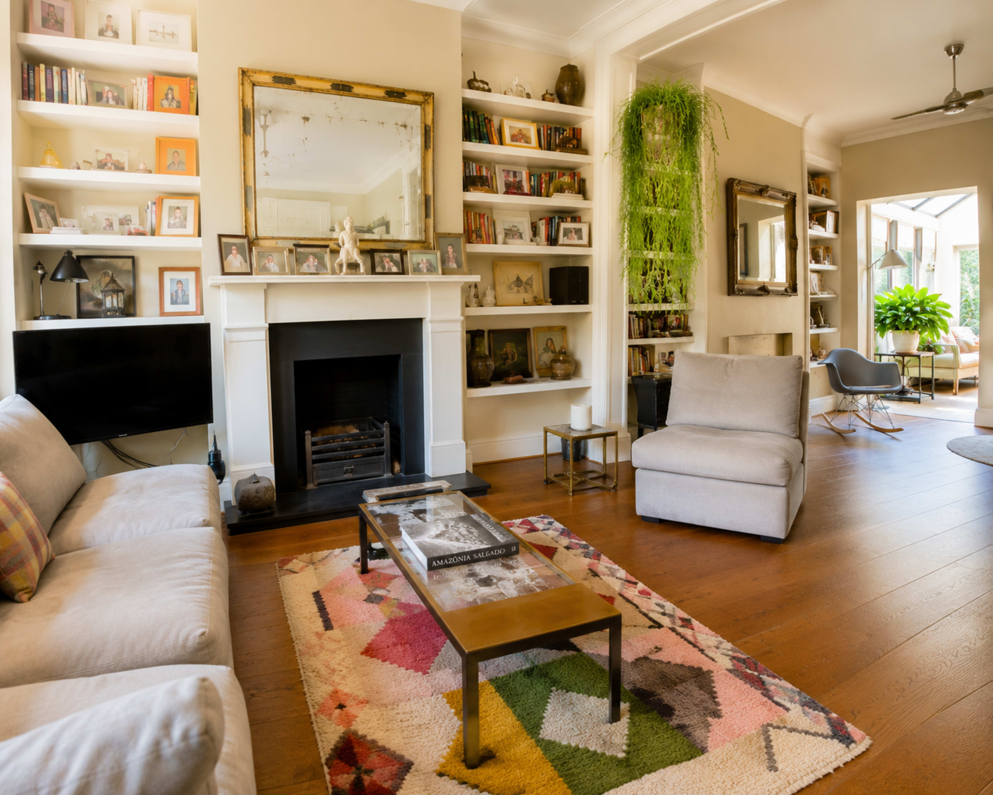 The living room features a fireplace, shelves filled with framed pictures, a coffee table, a large rug, and a view of a bright space with plants.