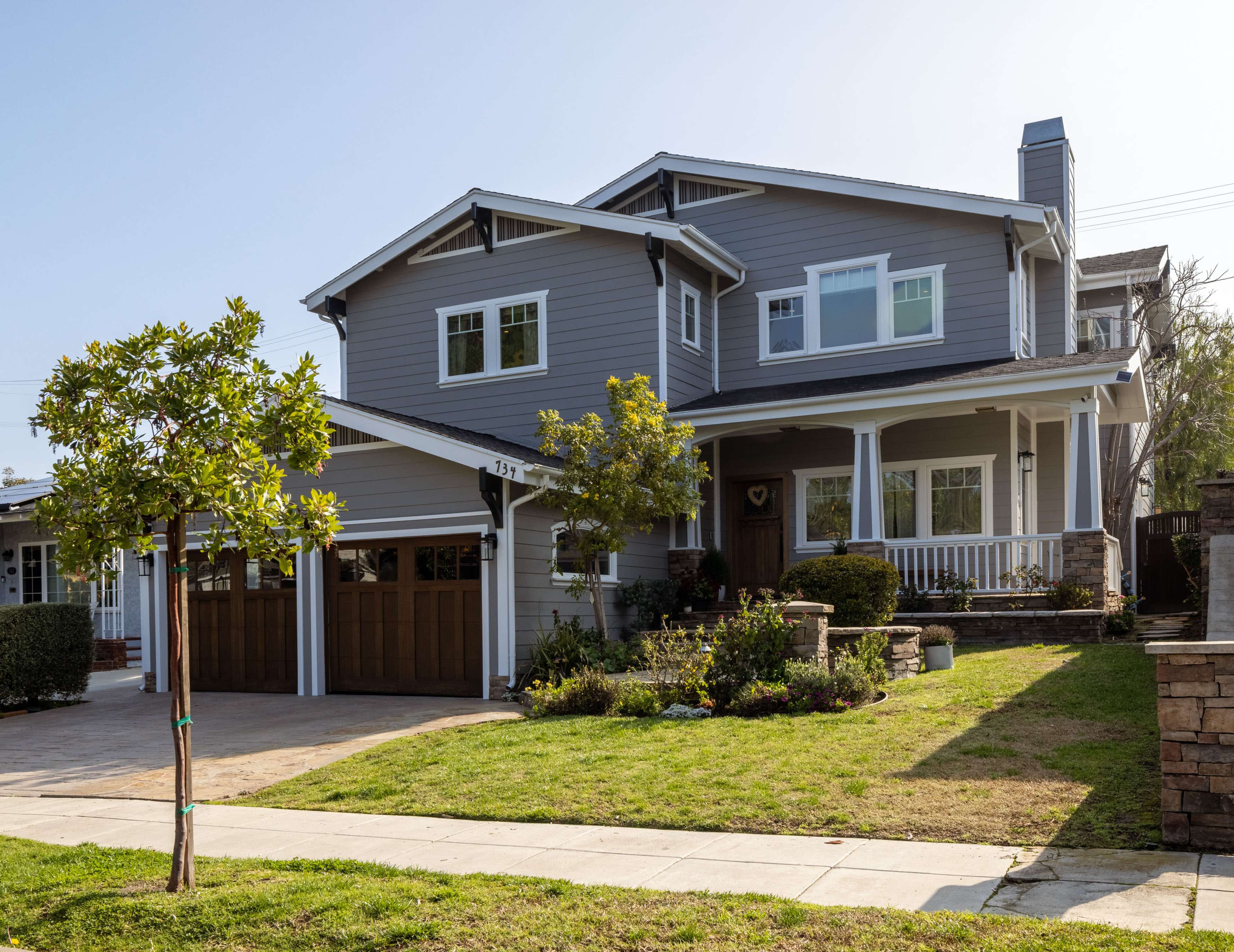 A two-story gray house with a wooden garage door and a front porch is situated on a landscaped yard.
