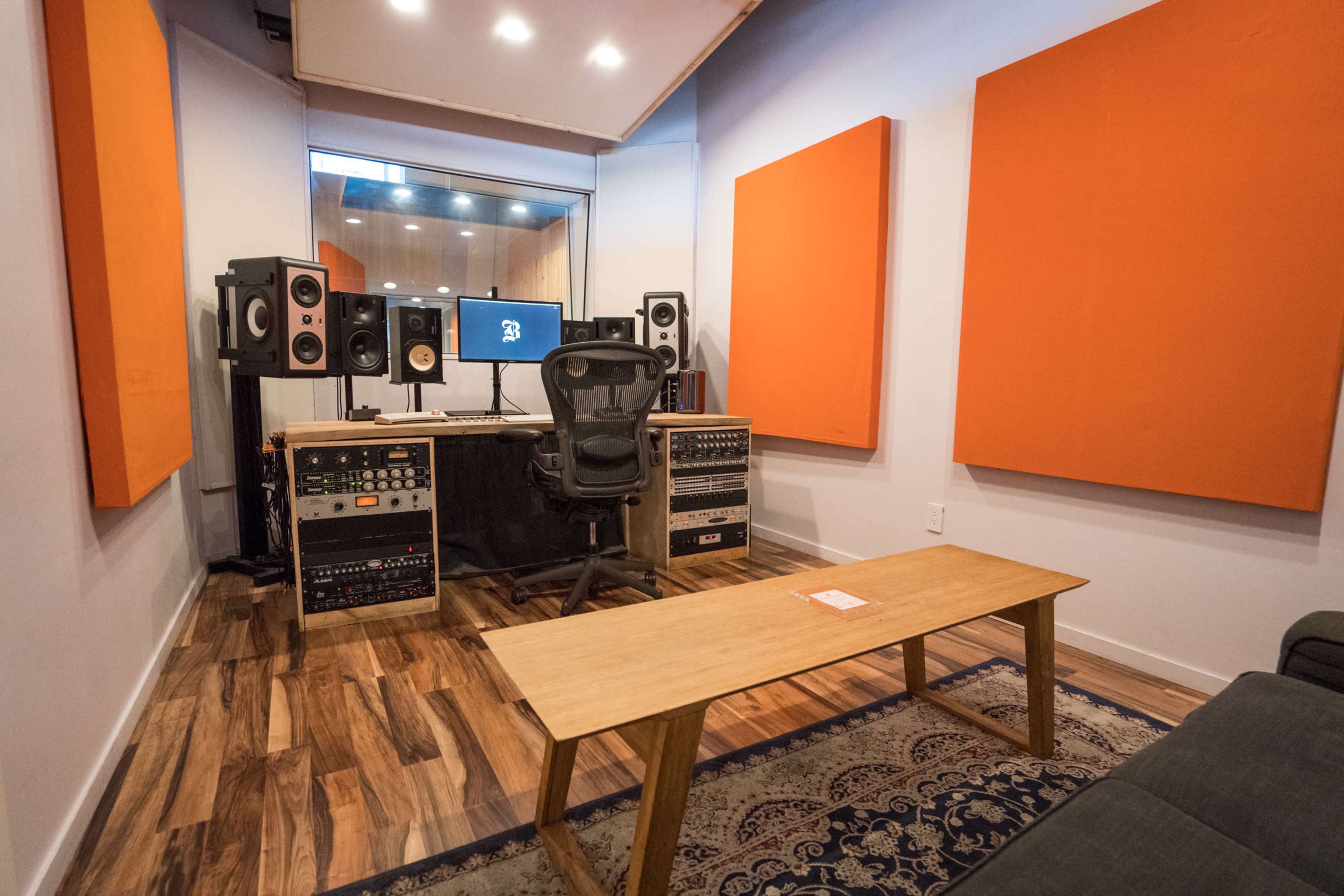 A soundproof recording studio with orange acoustic panels, a mixing desk, studio monitors, and a central chair facing computer screens.