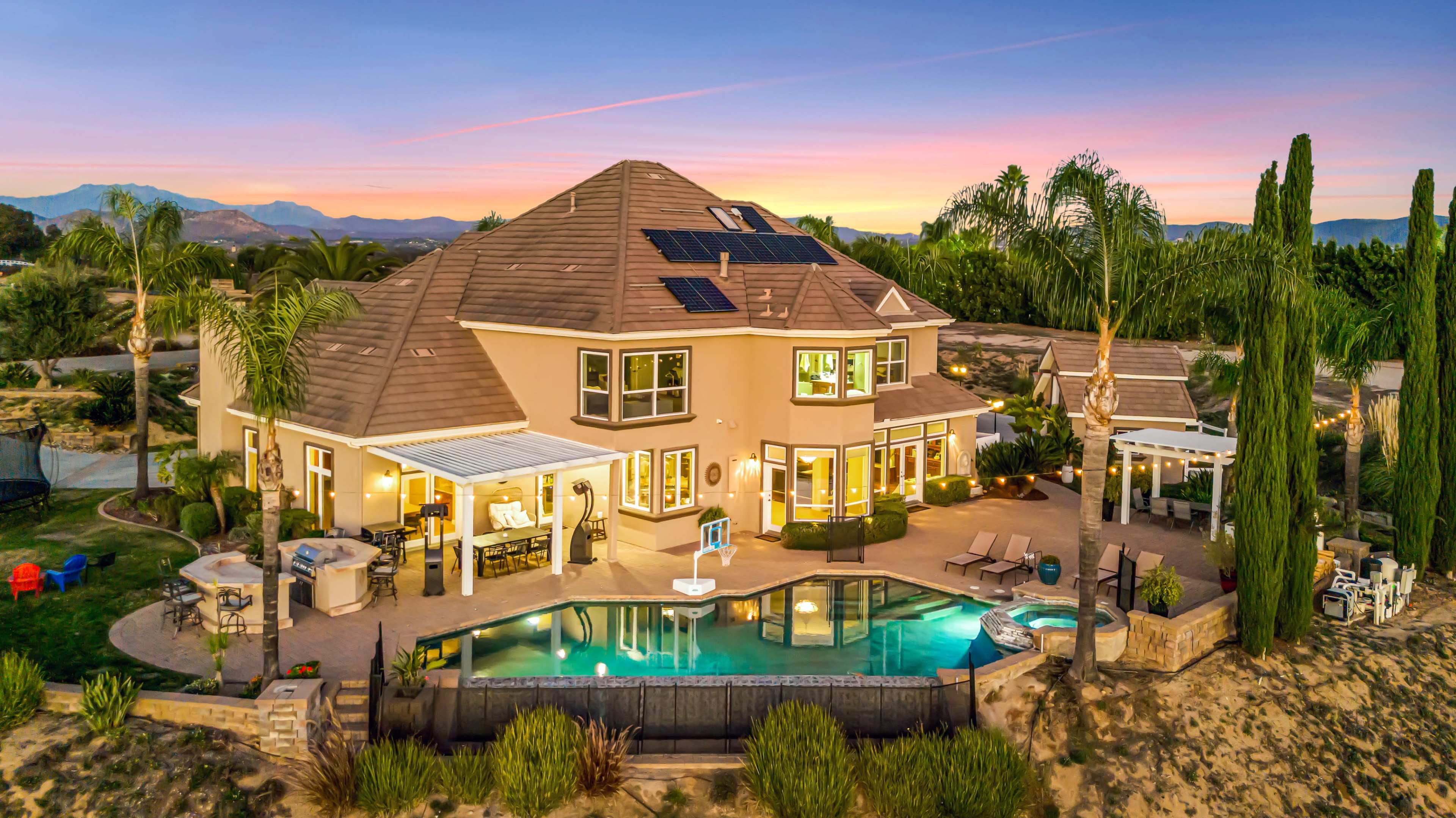 The image shows a large, two-story home with a pool and patio area, surrounded by palm trees and mountain views at sunset.
