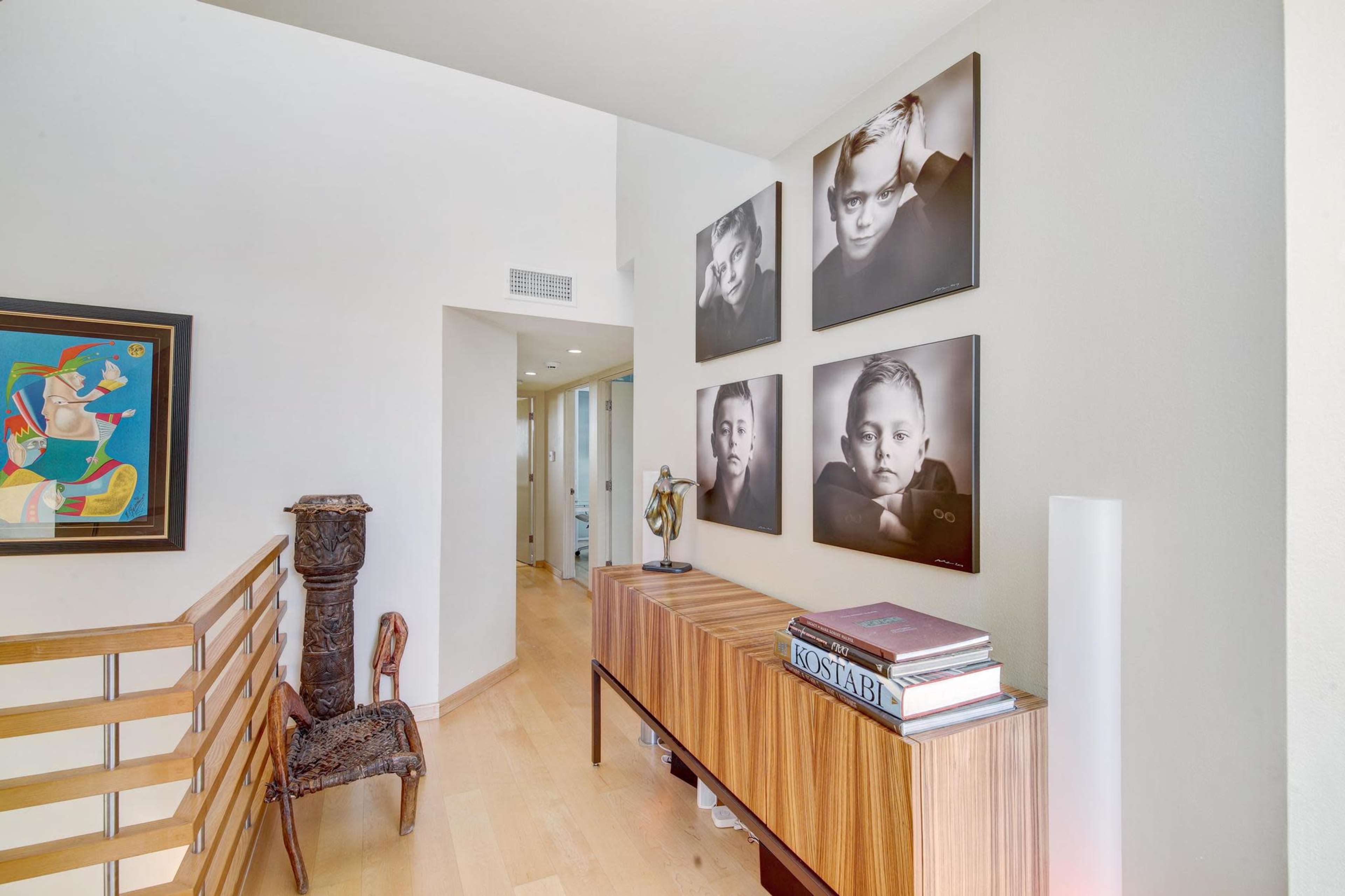 The image shows a hallway featuring framed black-and-white portraits on a wall beside a wooden cabinet with art books and a sculpture.