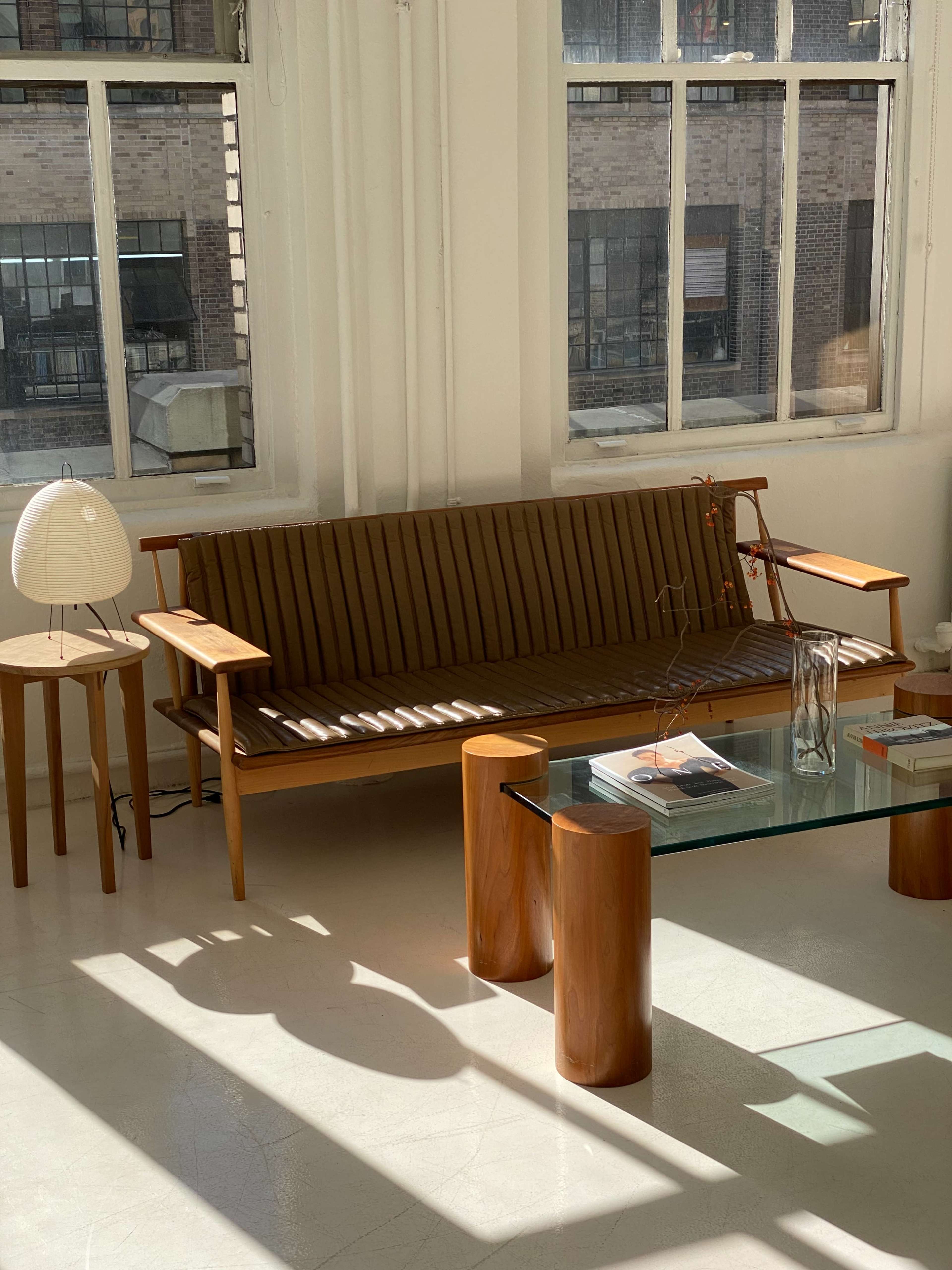A wooden slatted sofa is positioned in a brightly lit room with large windows, accompanied by a round side table and a glass coffee table.