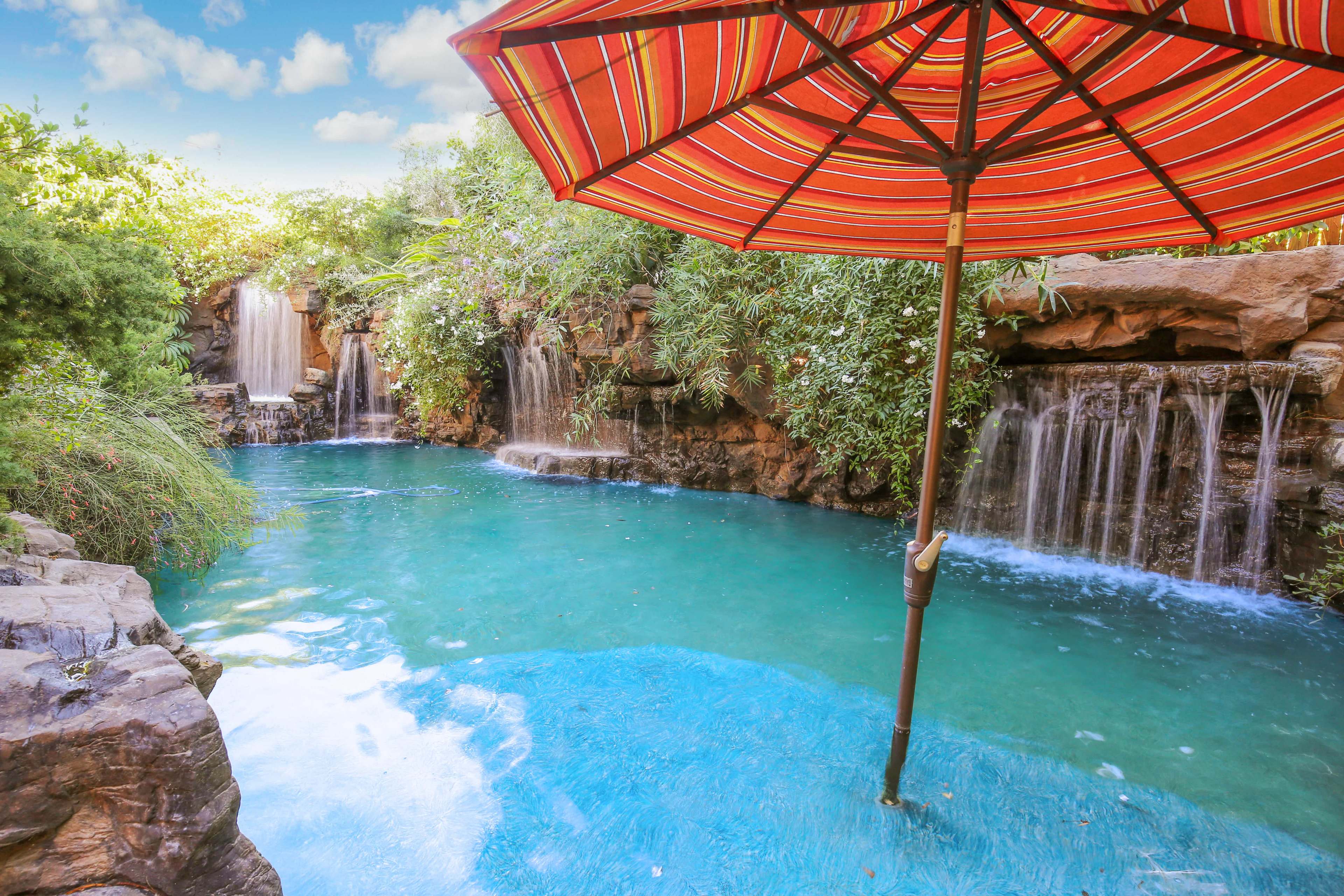 A vibrant pool area with a striped umbrella, surrounded by lush greenery and two small waterfalls cascading into the water.