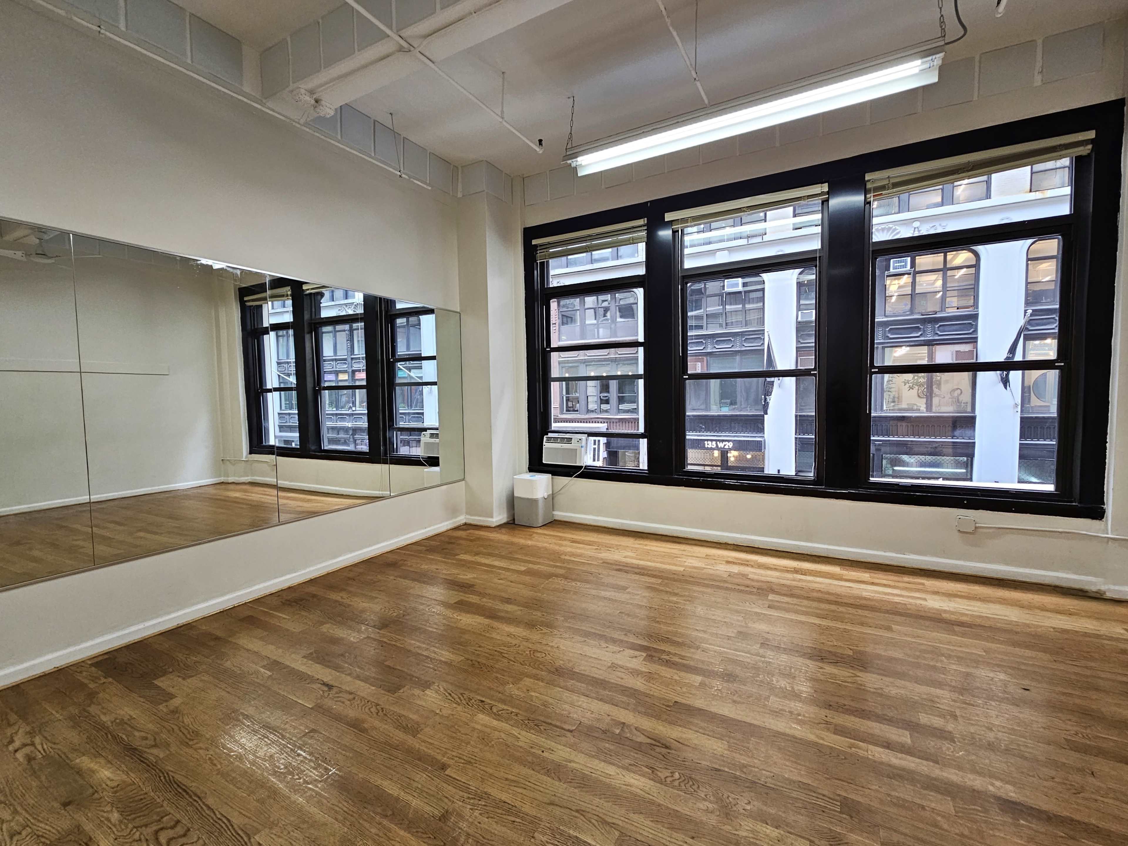 A spacious, empty room with hardwood floors, large windows, and a mirrored wall reflecting the street outside.