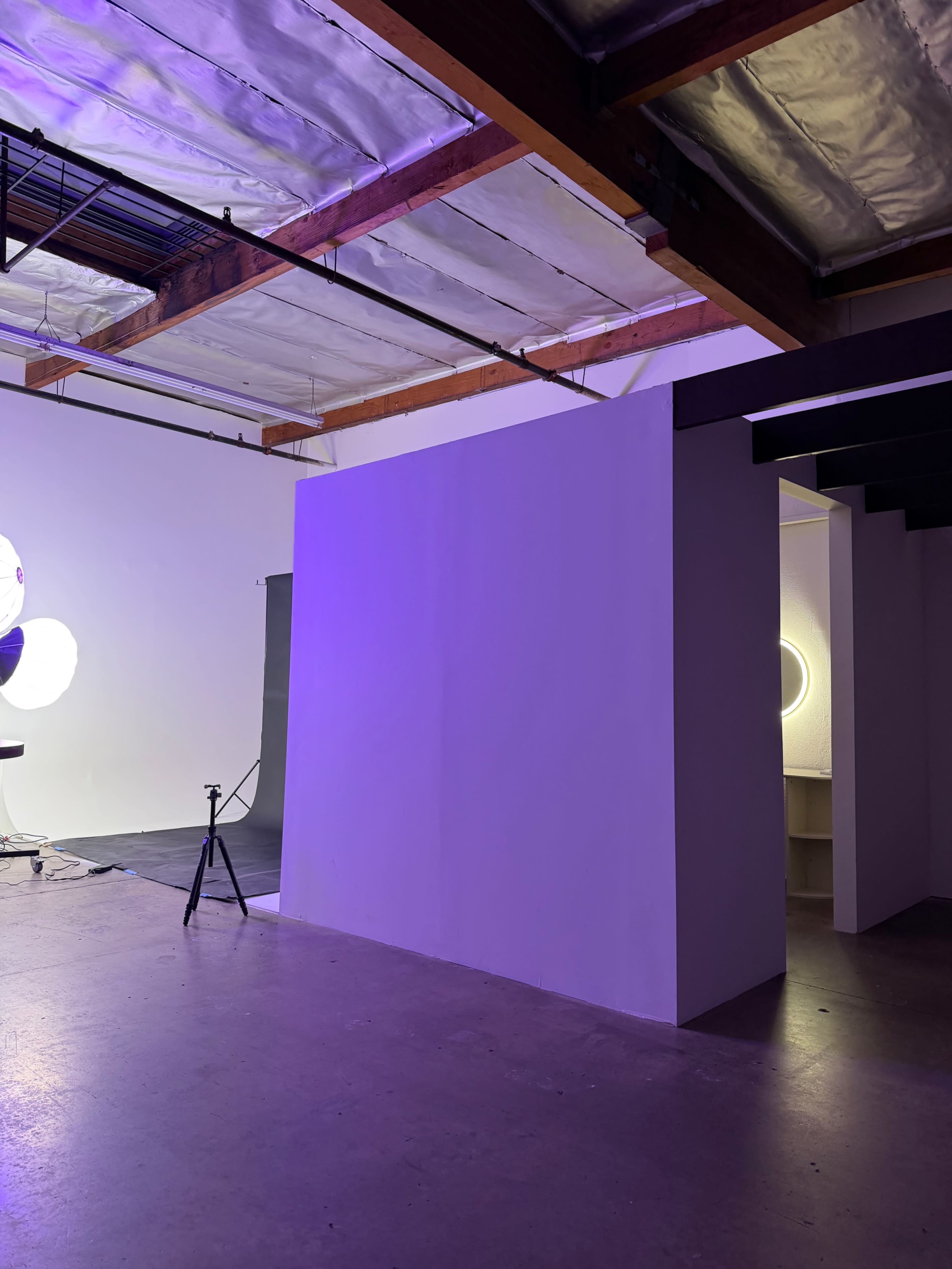 The image shows an indoor space with a white wall, a circular light source, and a tripod setup on a concrete floor.
