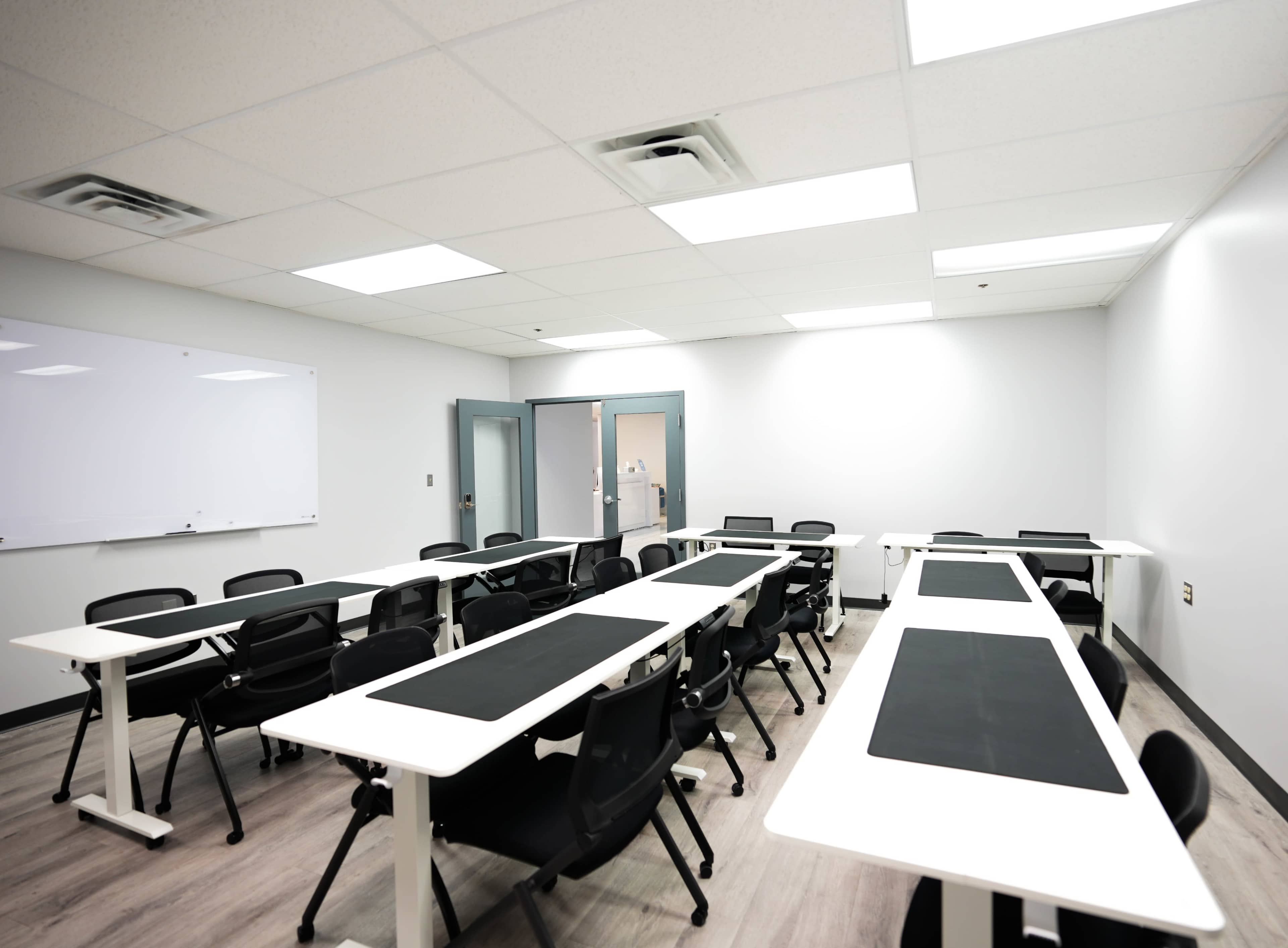 The image shows a modern classroom with several white desks arranged in rows, black chairs, and a whiteboard on one wall.