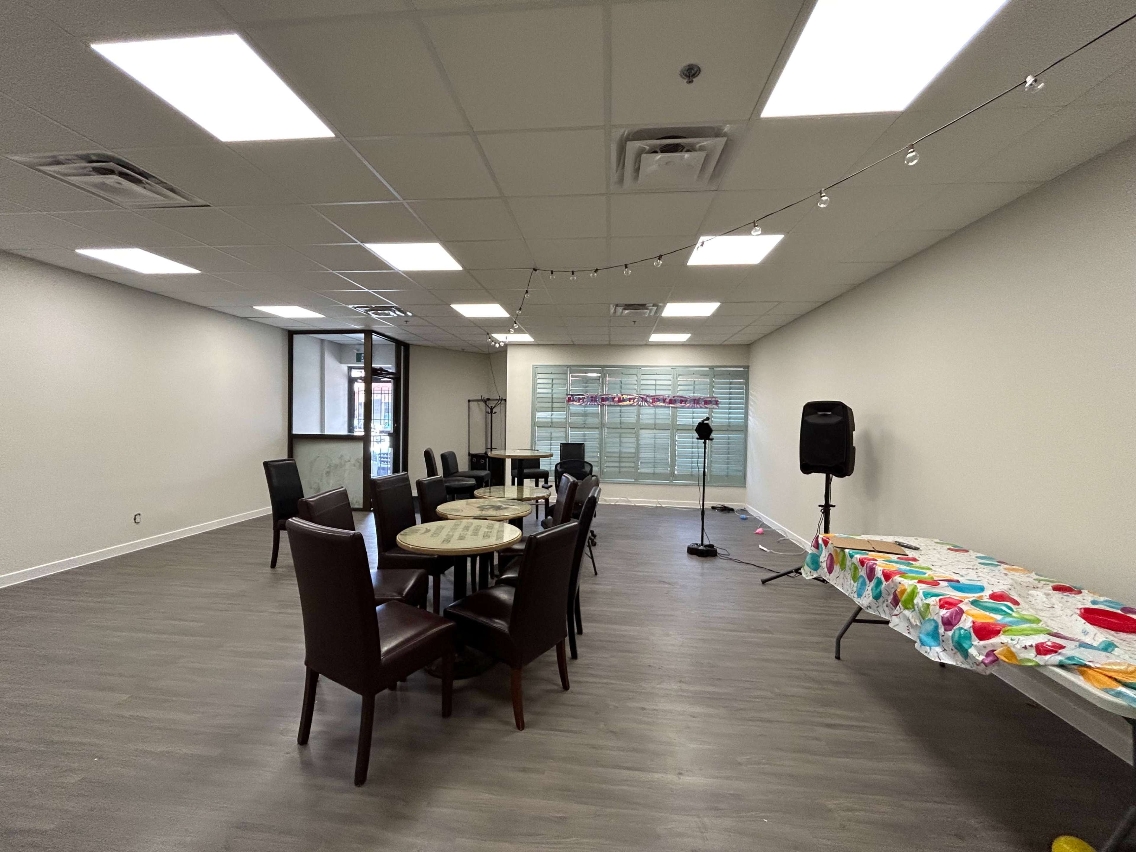 The image shows a spacious indoor area featuring several tables and chairs, along with a speaker setup and a table covered with a colorful cloth.