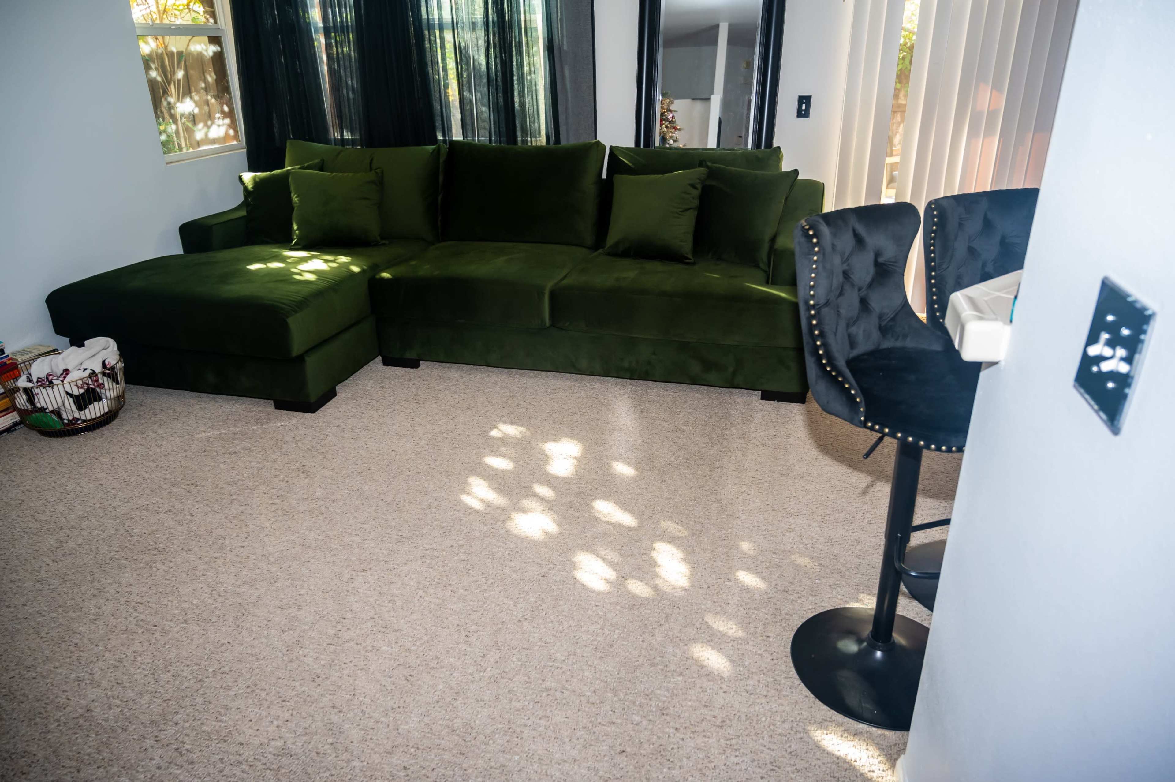 A green sectional couch with cushions in a living room with light streaming through the window onto a beige carpet.