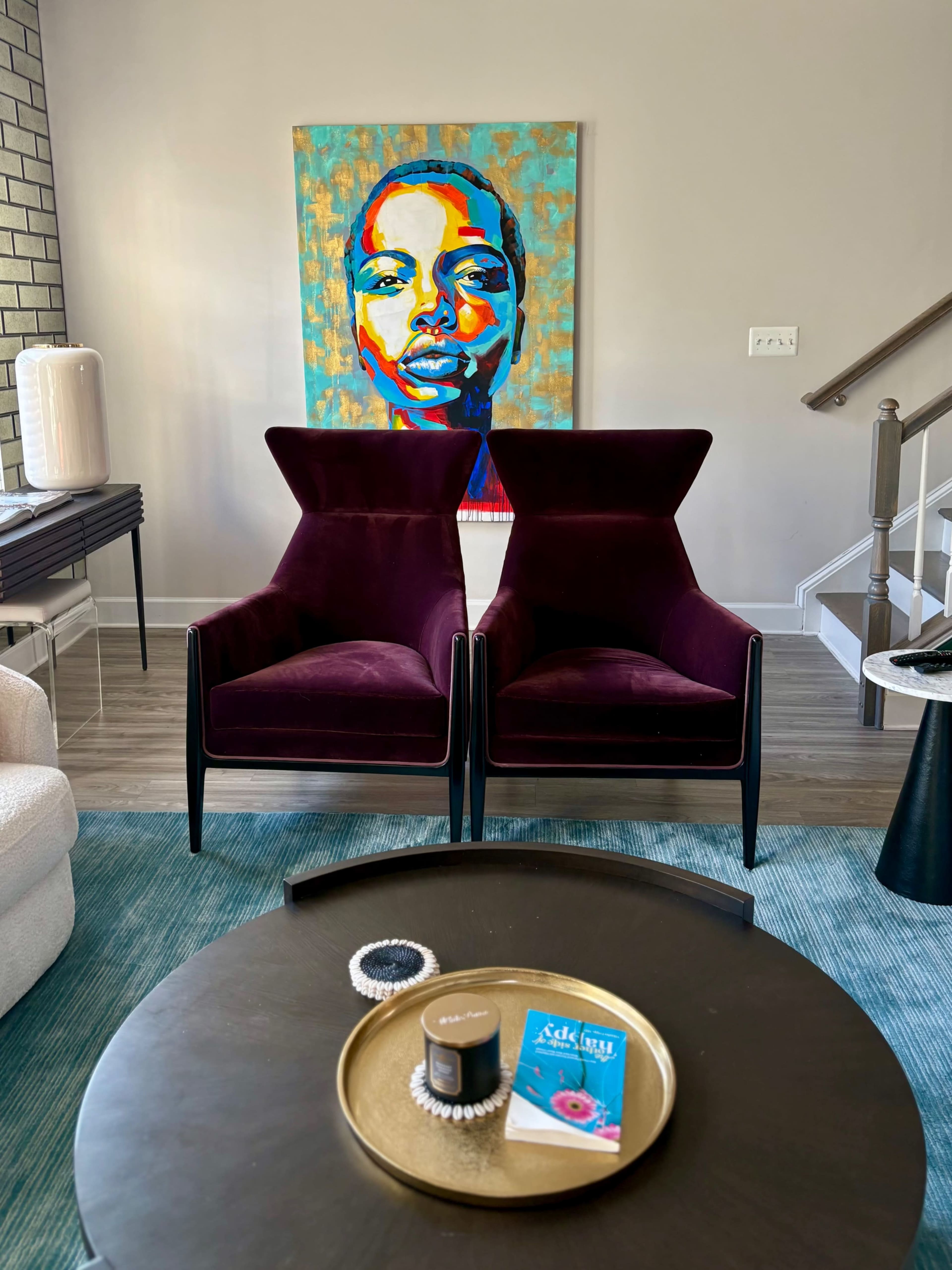 The image shows two purple chairs facing each other in a living room, with a colorful portrait of a person on the wall behind them.