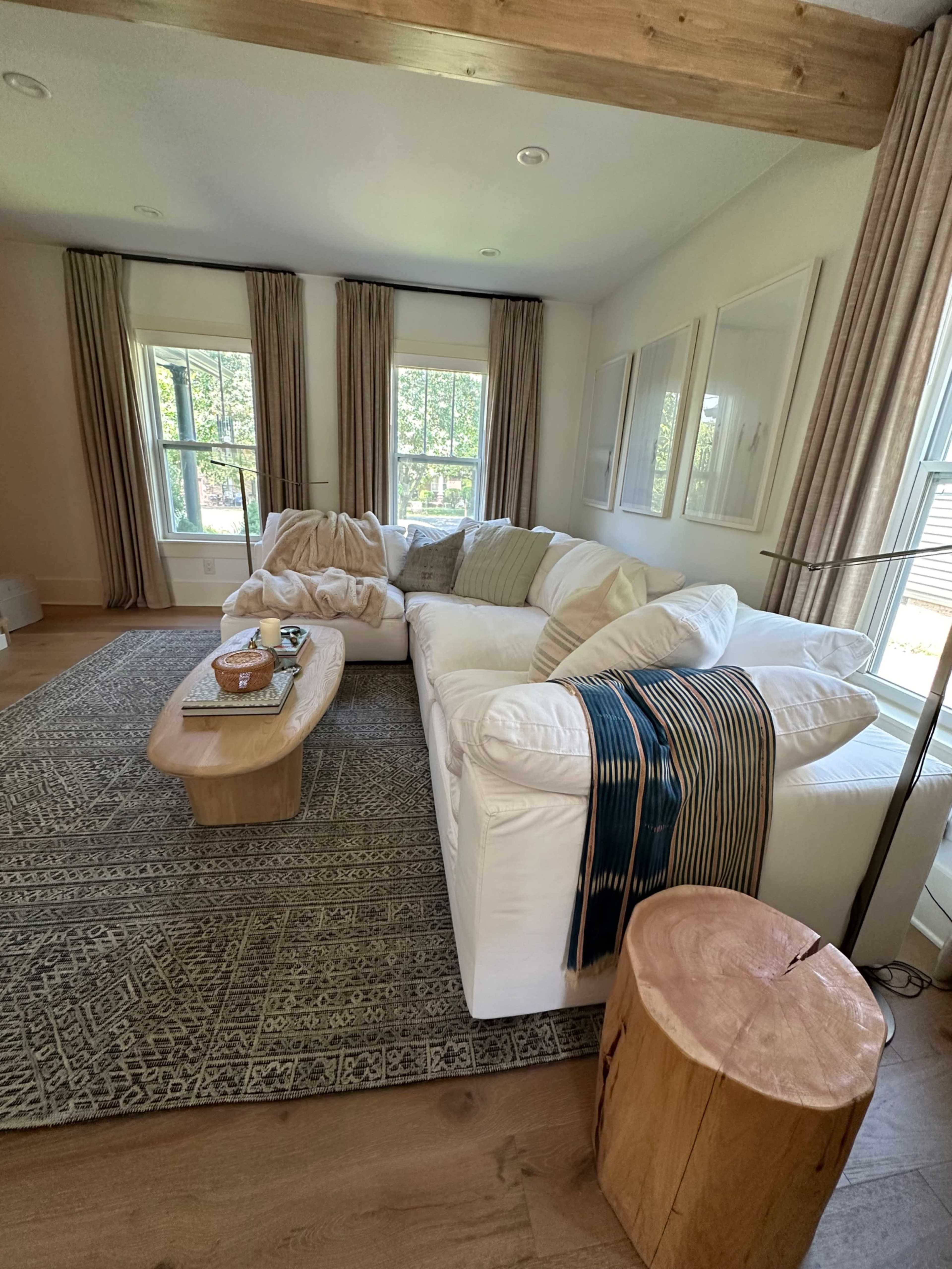 A bright living room features a large white sectional sofa, a wooden coffee table, and two tall windows adorned with curtain panels.
