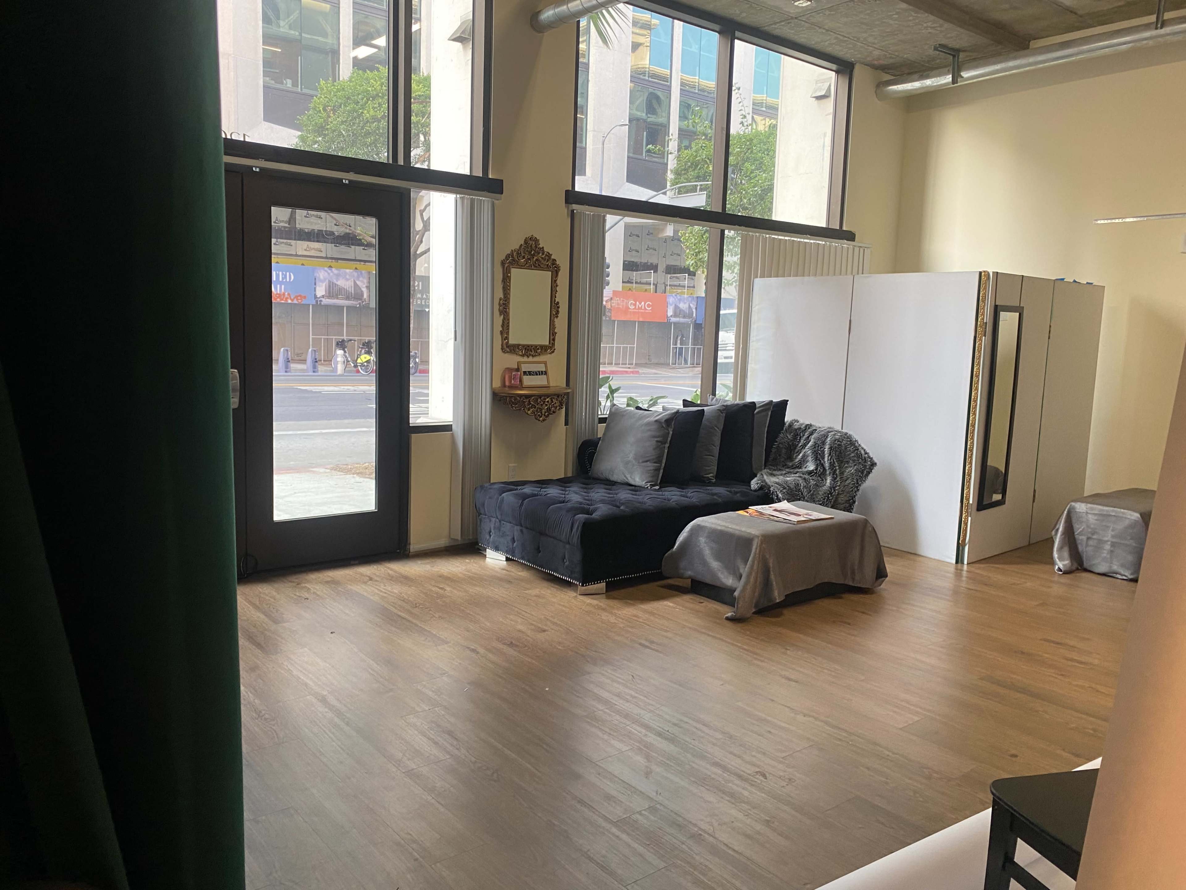 The image shows a stylish interior space with a dark couch, a large mirror, and a partition, all illuminated by natural light from large windows.