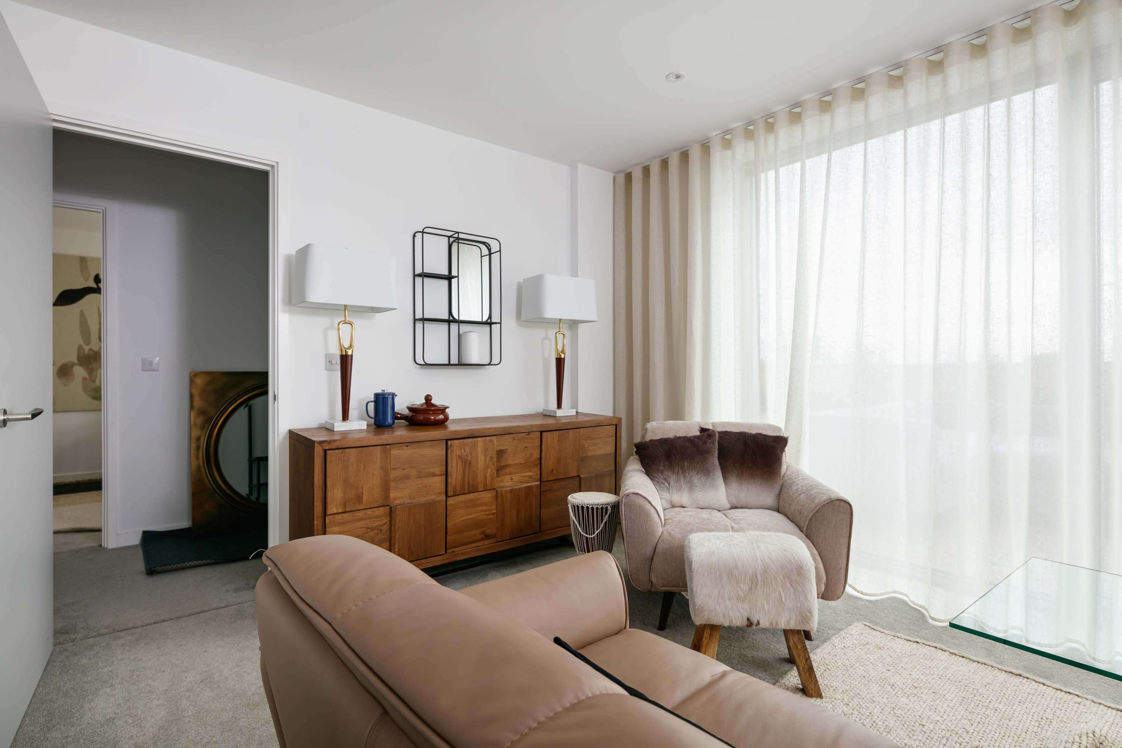 A modern living room features a wooden sideboard, soft furniture, and large sheer curtains allowing natural light to filter in.