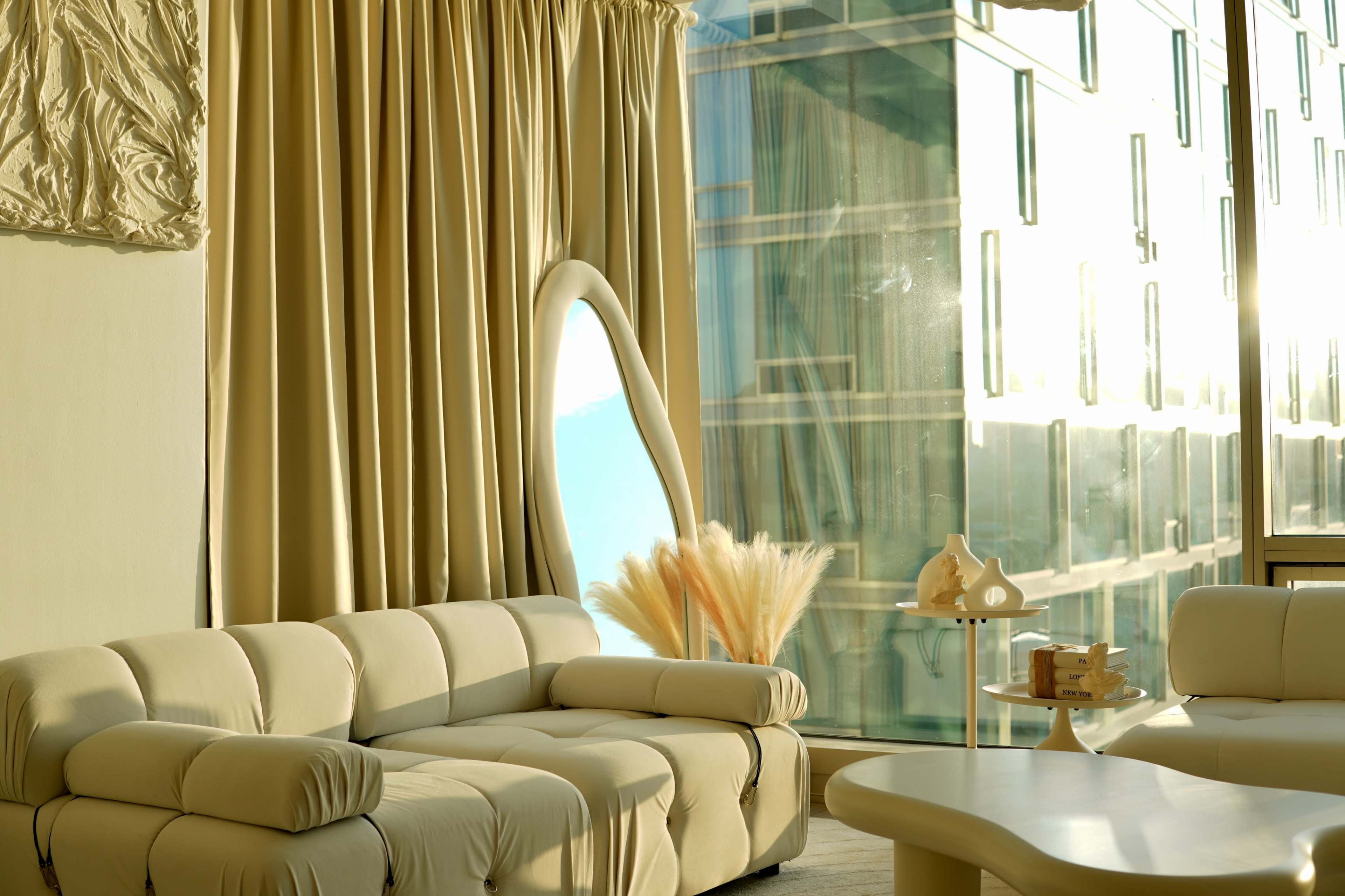 A modern living room features a light-colored couch, large curtains, and a decorative arrangement of dried plants beside a floor-to-ceiling window showing a view of an urban exterior.
