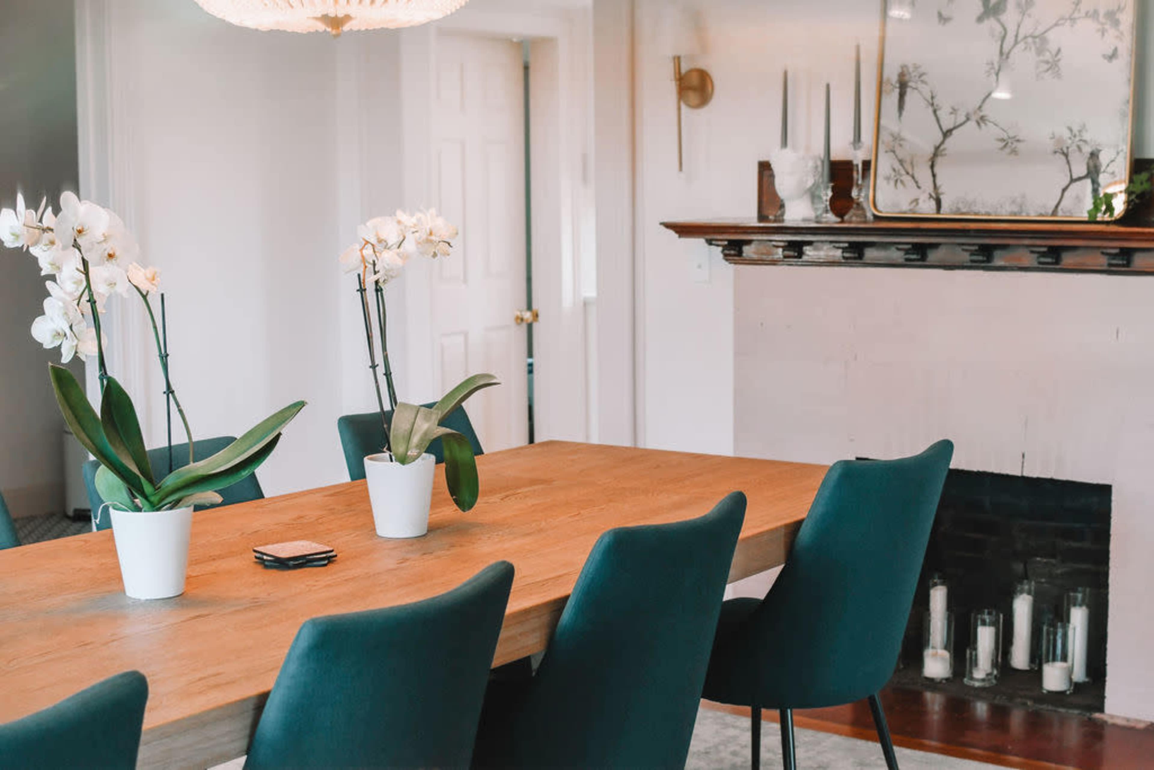 A wooden dining table with green chairs is set in a well-lit room featuring potted orchids and a decorative fireplace.
