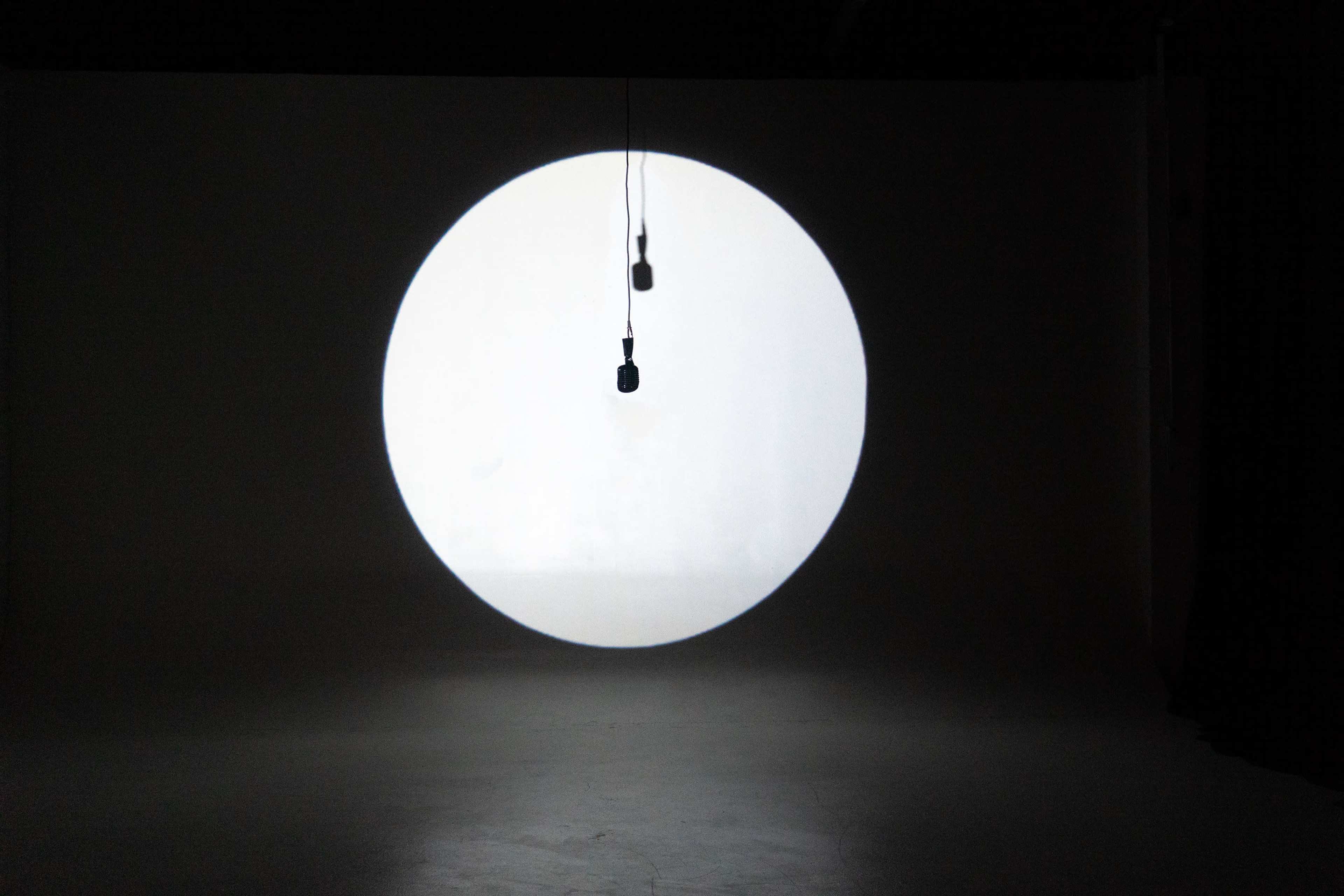 A single microphone hangs in front of a large, illuminated circular background in a dimly lit room.