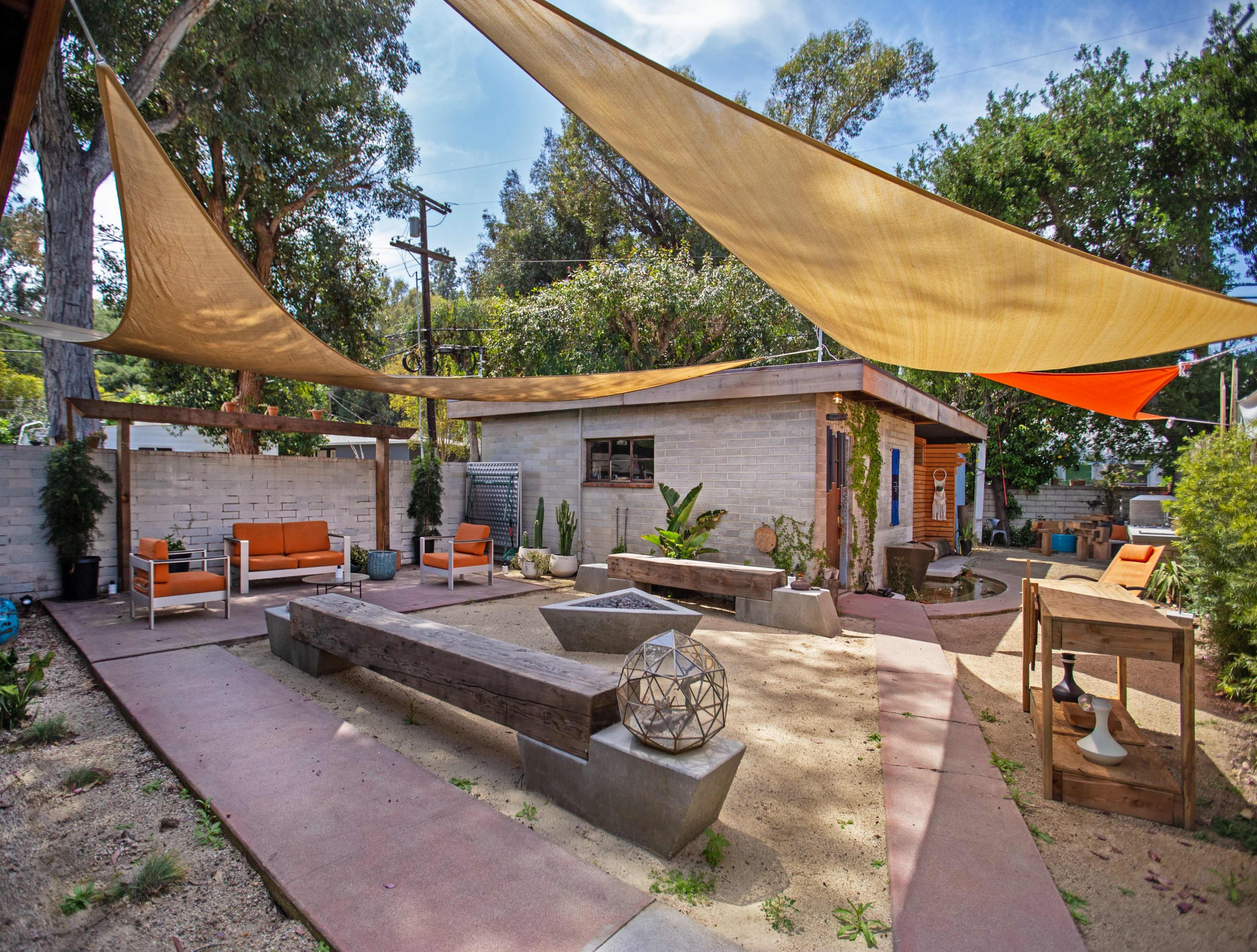 A landscaped outdoor space featuring sandy areas, seating arrangements under large fabric canopies, and a central fire pit surrounded by wooden benches.