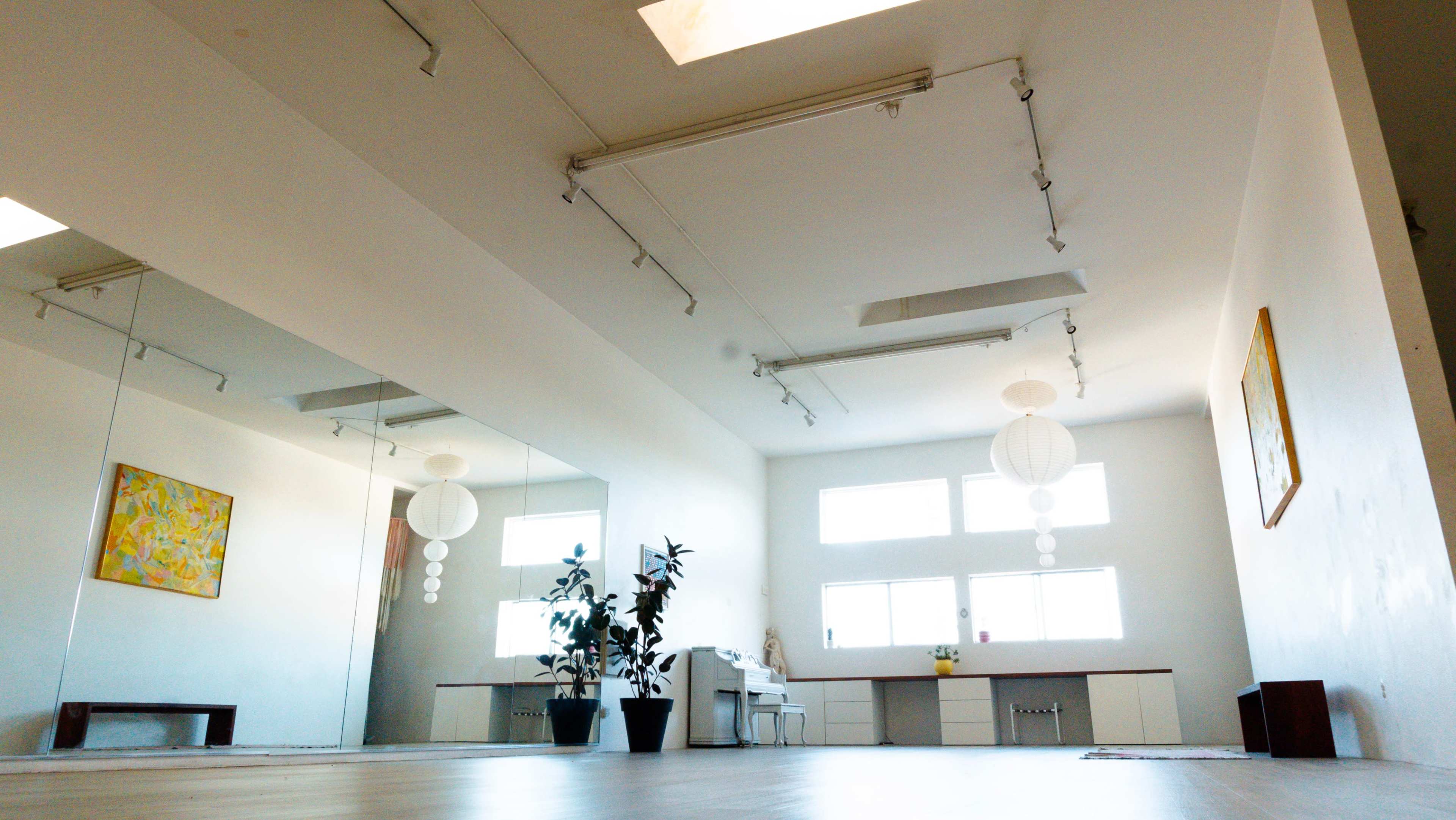 The image shows a spacious room with mirrored walls, natural light from large windows, and a few decorative elements including plants and hanging light fixtures.