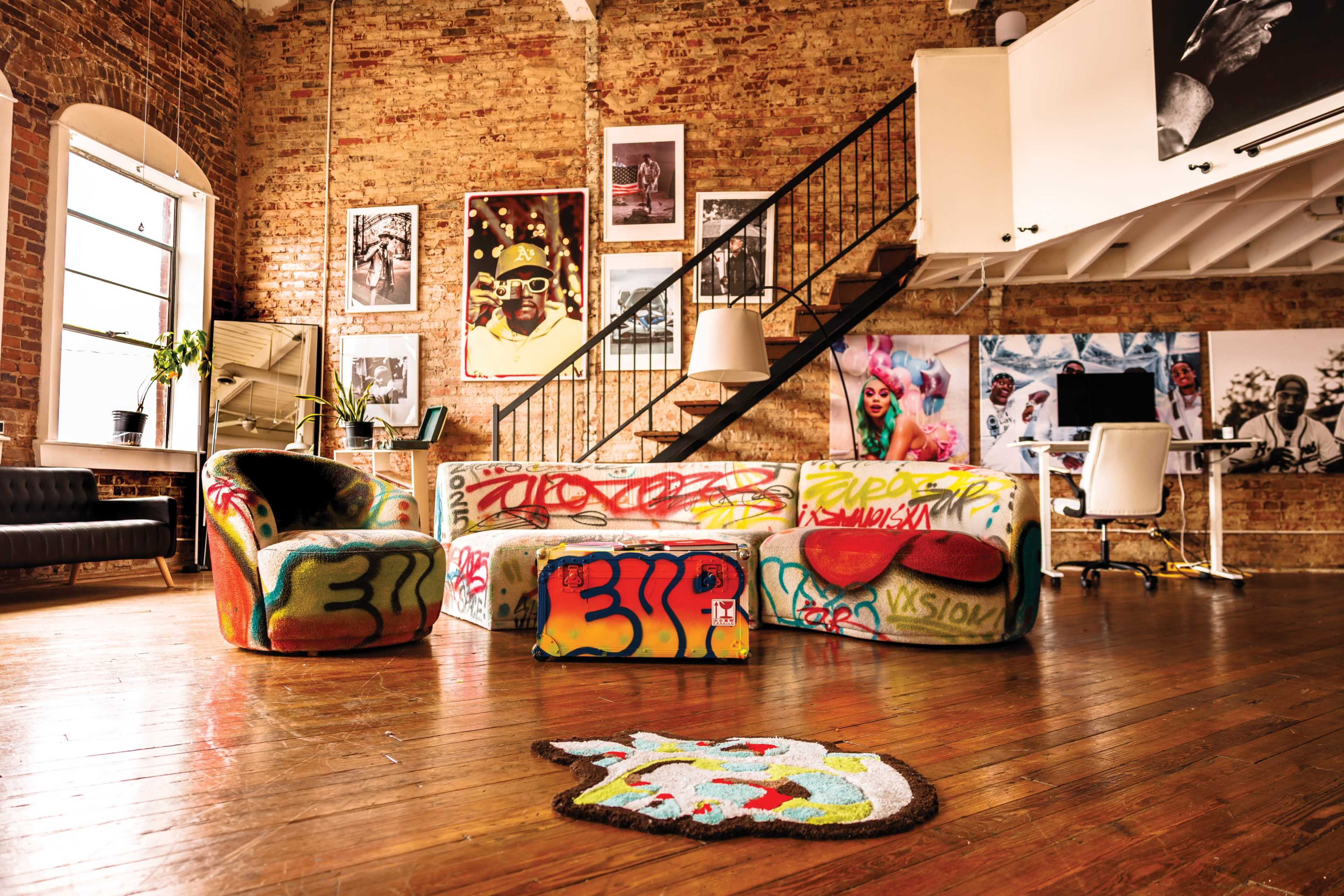 A spacious loft features a colorful, graffiti-covered couch and ottoman, wooden floors, and a staircase leading to an upper area with a workspace.