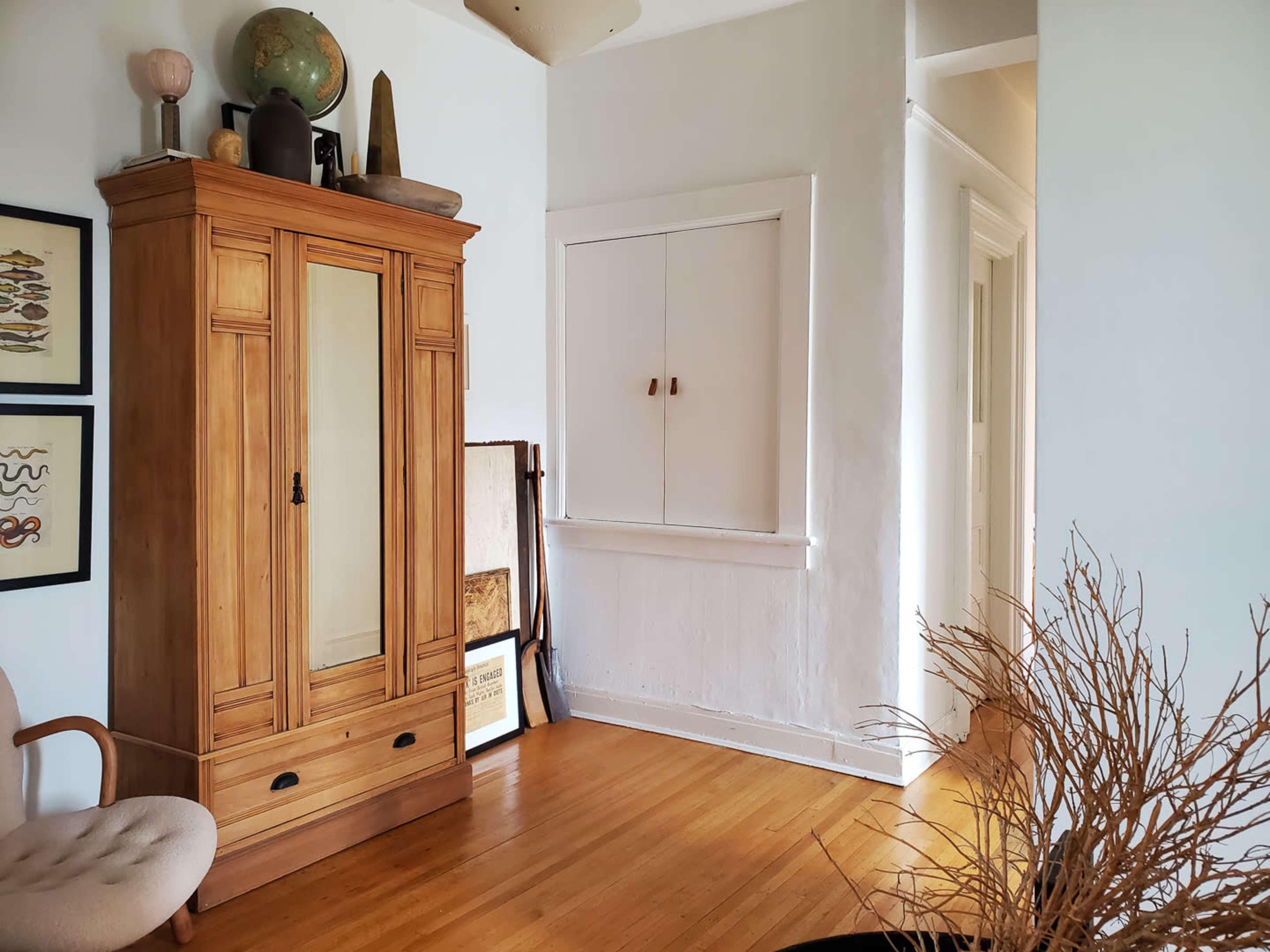 The image shows a room with wooden flooring featuring a tall, light-colored armoire, a beige chair, and a decorative plant in the corner.
