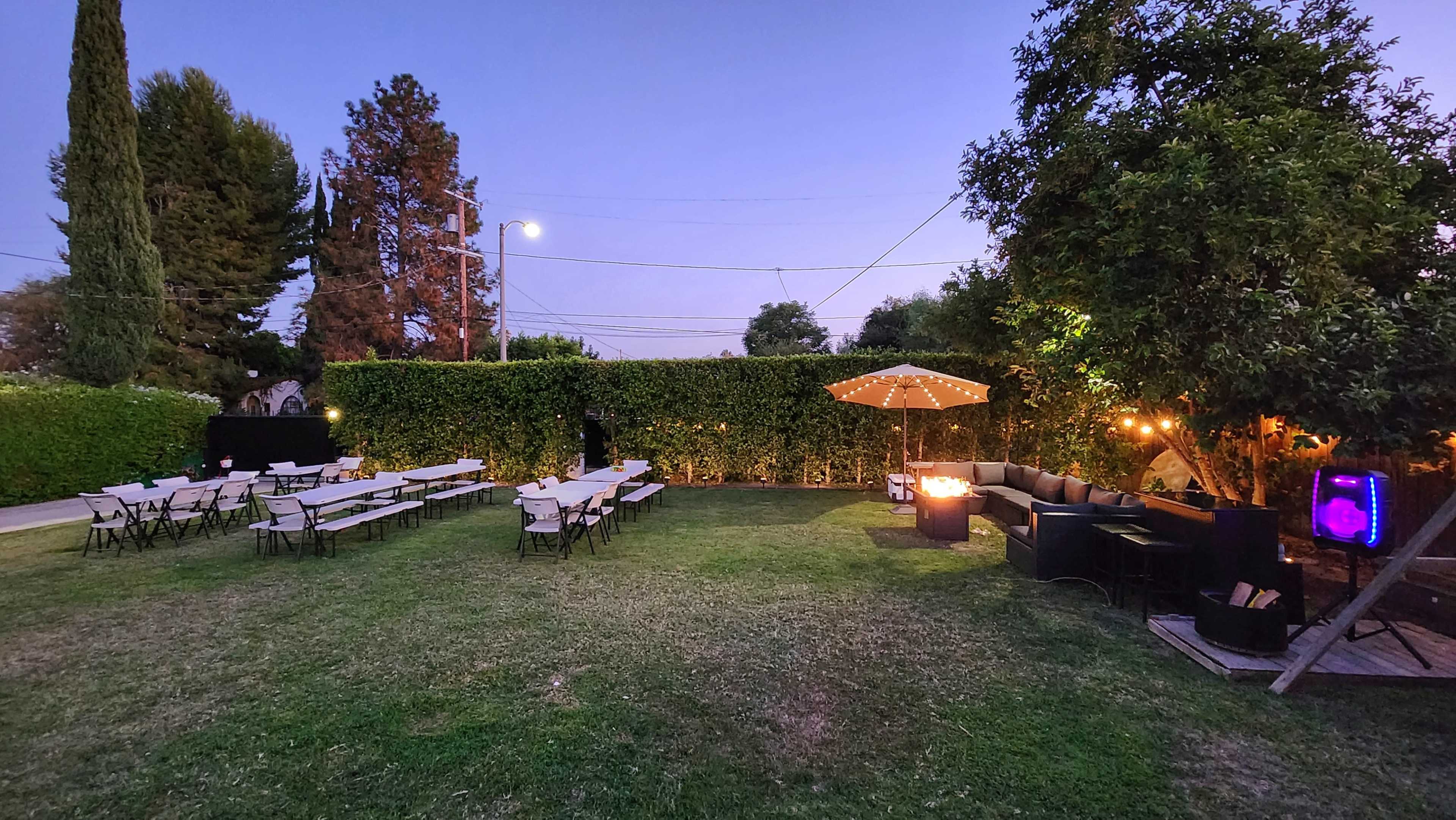 The image shows a backyard setting with rows of chairs and tables on a lawn, surrounded by tall hedges and trees, illuminated by string lights and a lamp post at dusk.