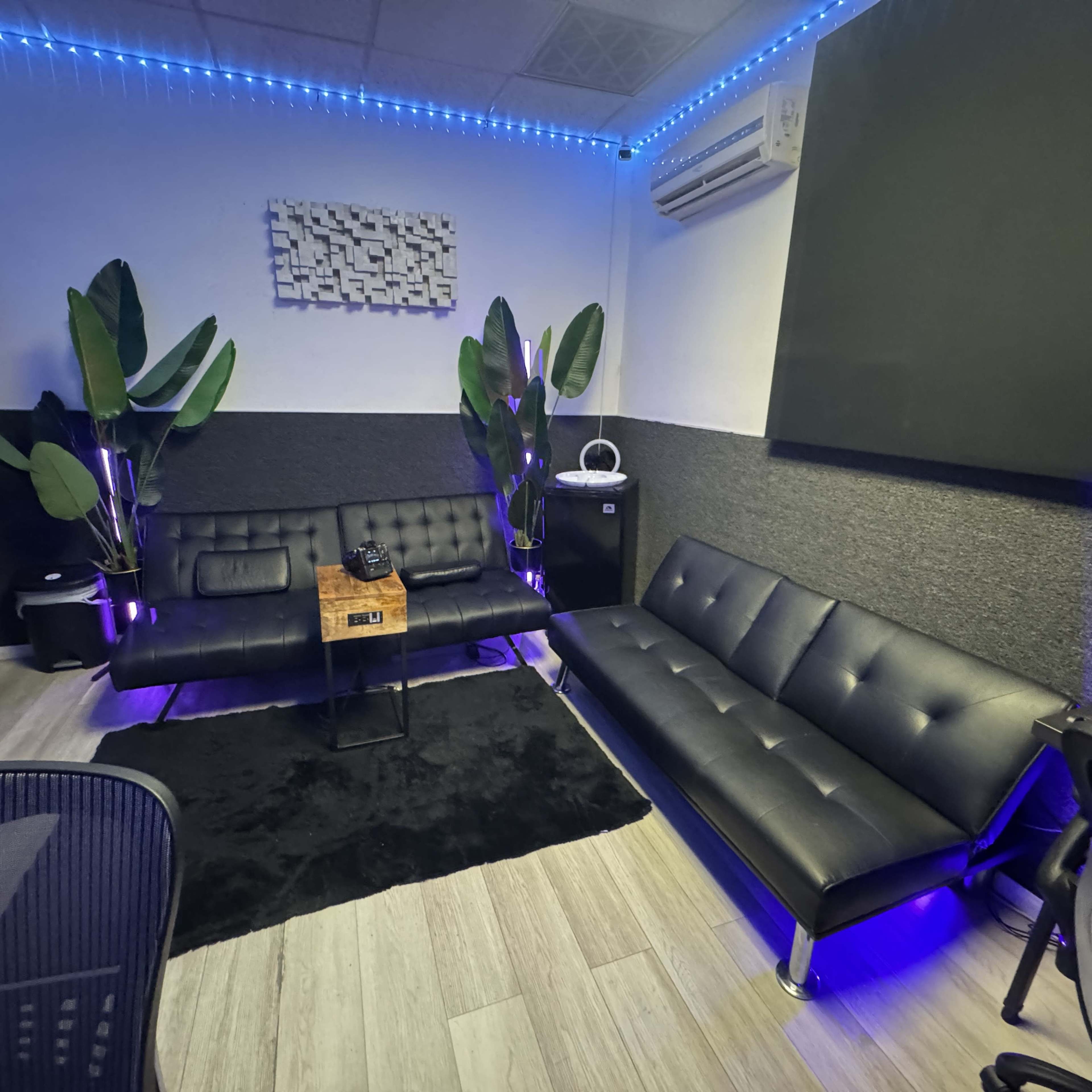 The image shows a modern lounge area with black sofas, a wooden side table, and plants, illuminated by blue LED lights.