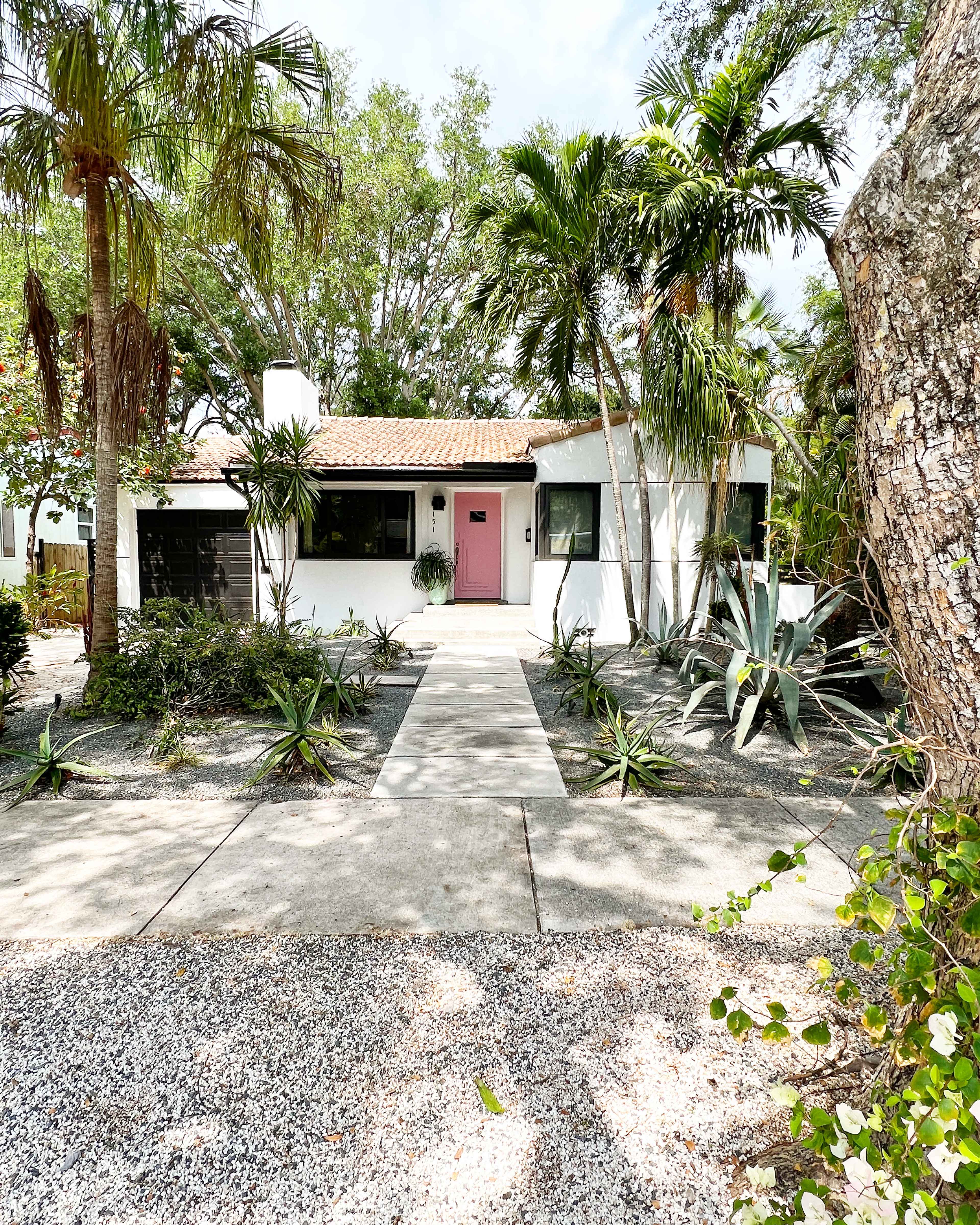 A single-story white house with a pink door is surrounded by tropical plants and a gravel pathway.