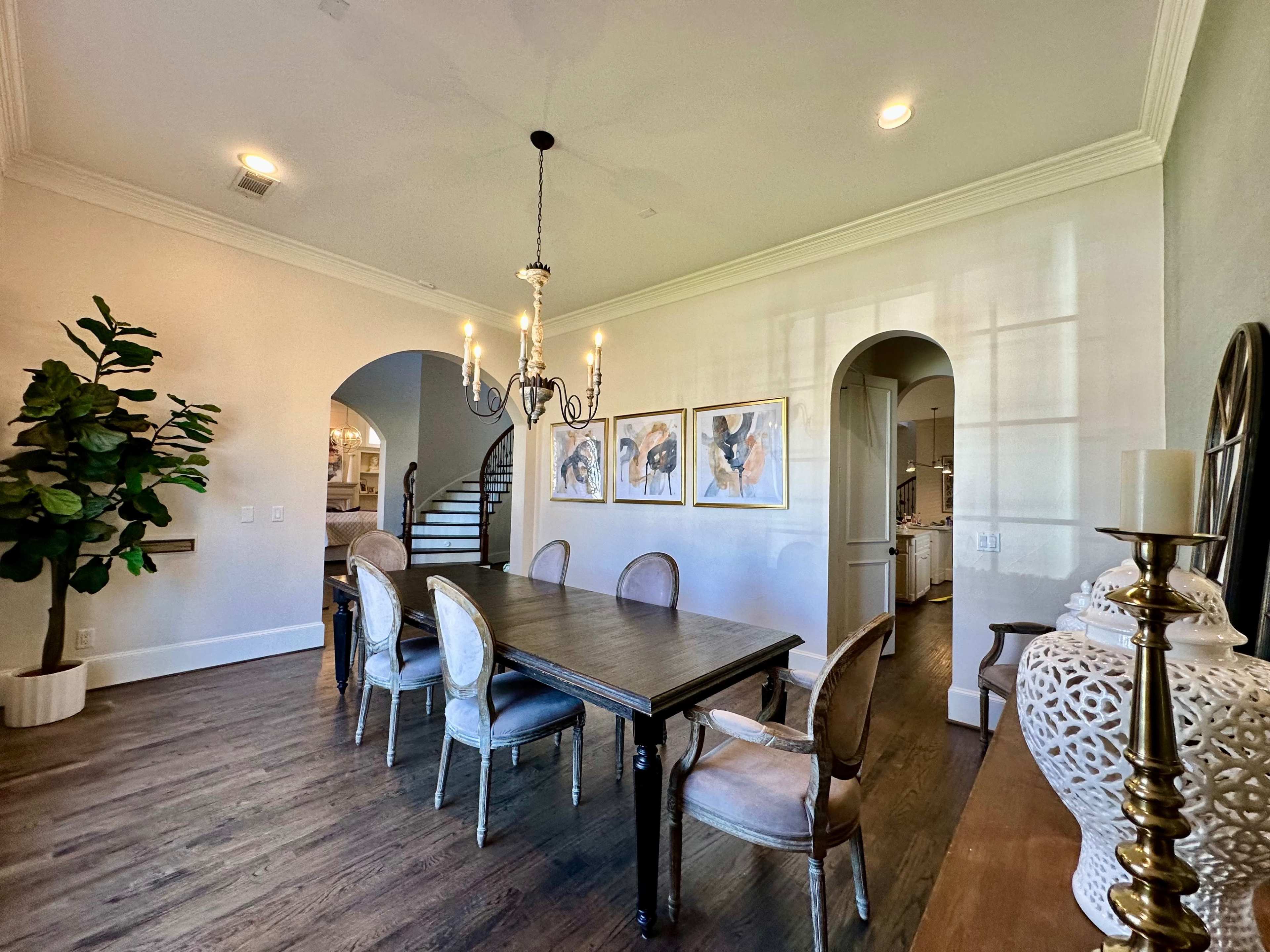 The image shows a dining room featuring a wooden table surrounded by upholstered chairs, with a chandelier hanging above and arched doorways leading to adjacent spaces.