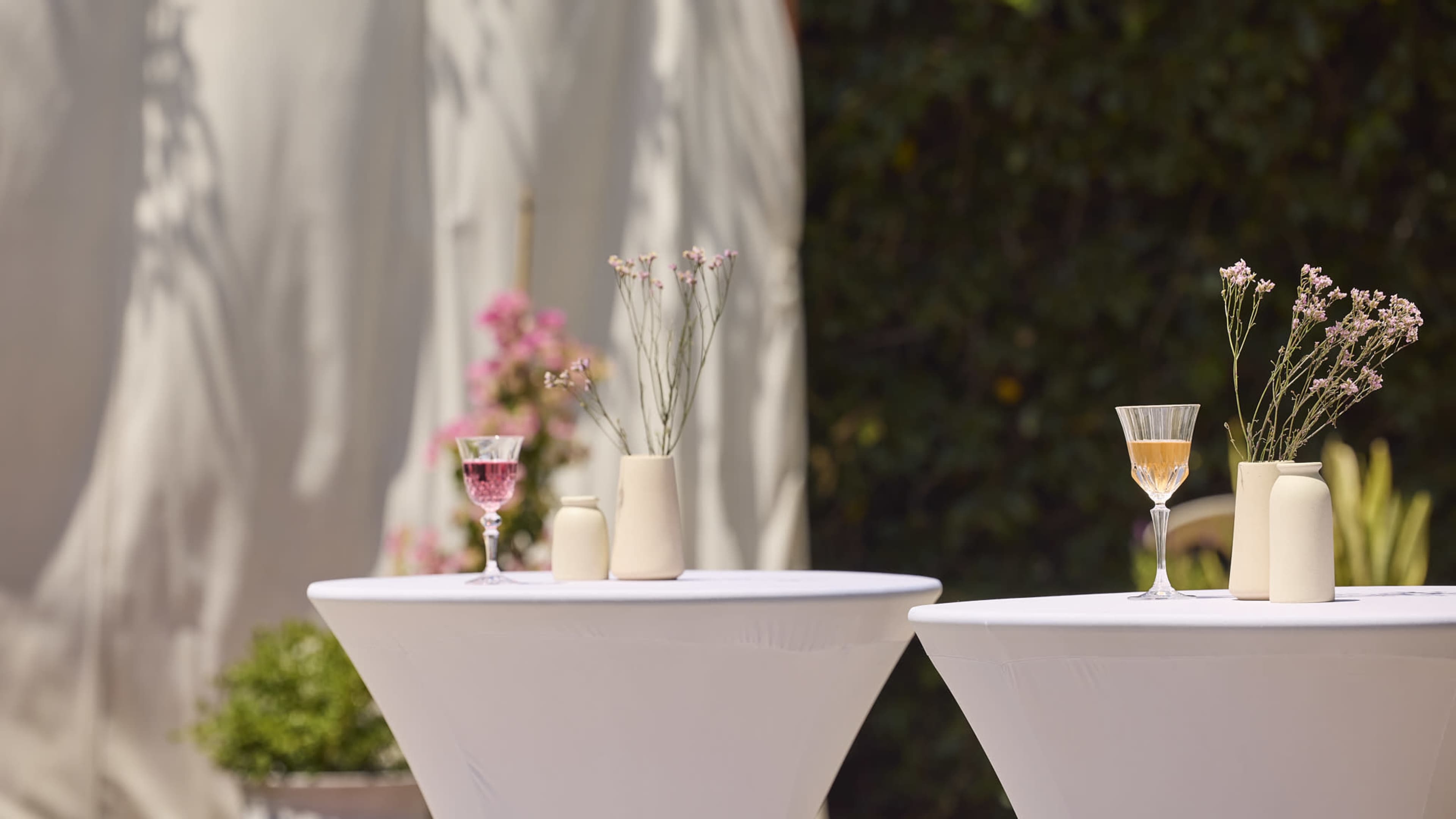Two round tables with white tablecloths are set outdoors, each featuring a glass of colorful drinks and small vases with flowers.