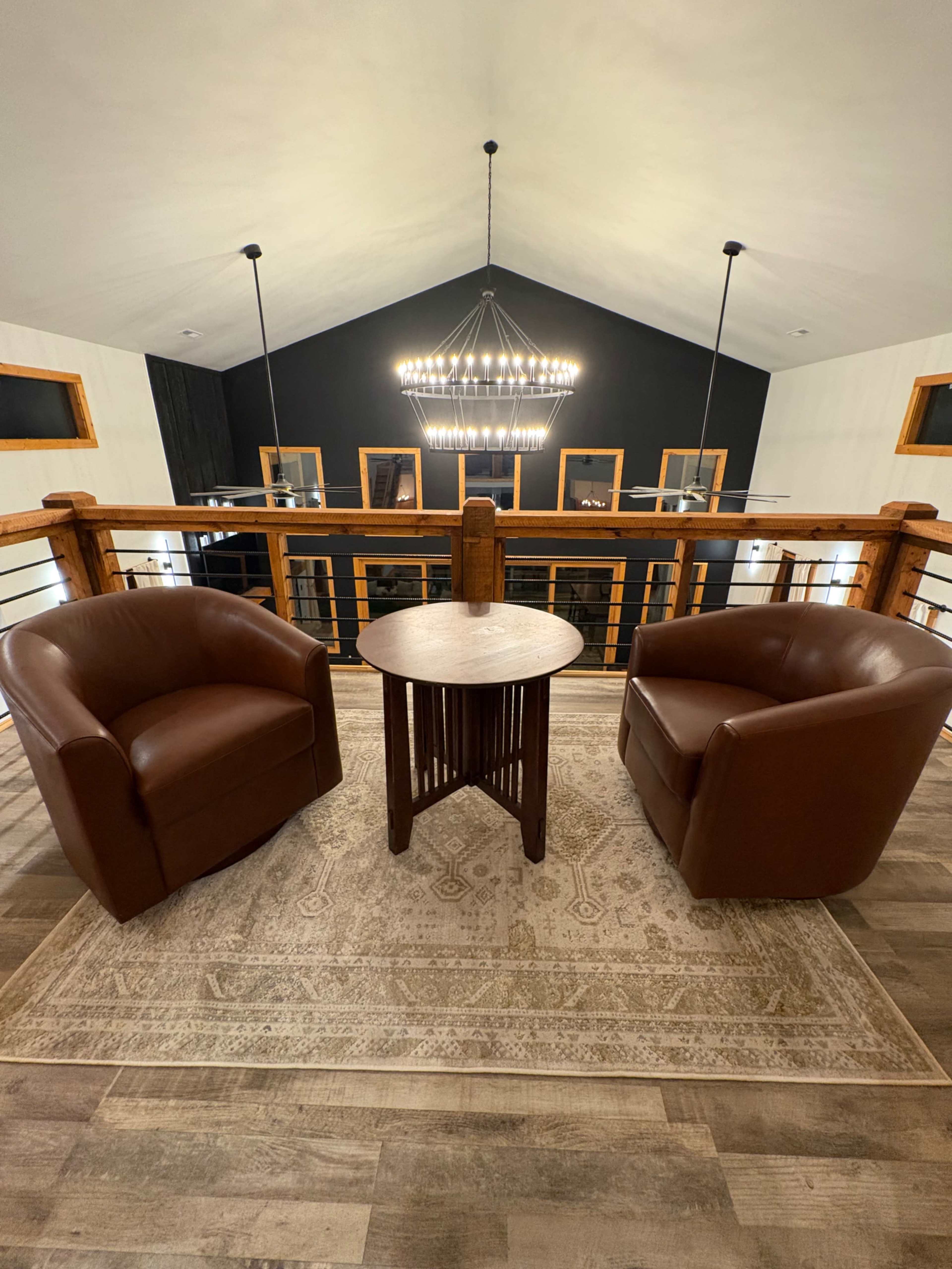 The image shows two brown armchairs facing each other across a round wooden table on a patterned rug, with a chandelier and framed pictures visible in the background.