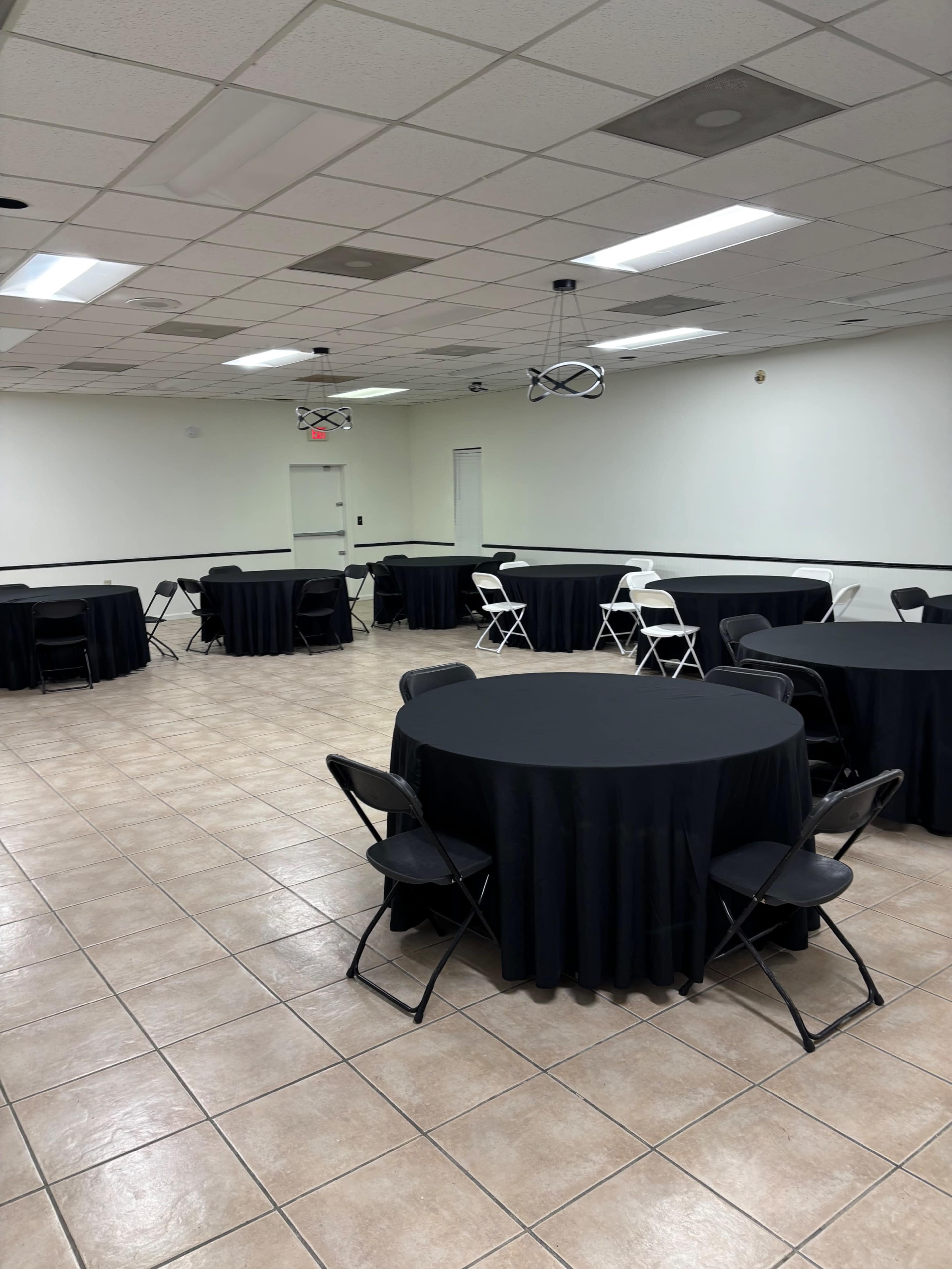 The image shows a spacious room with several round tables covered in black tablecloths, each accompanied by black and white folding chairs arranged around them.