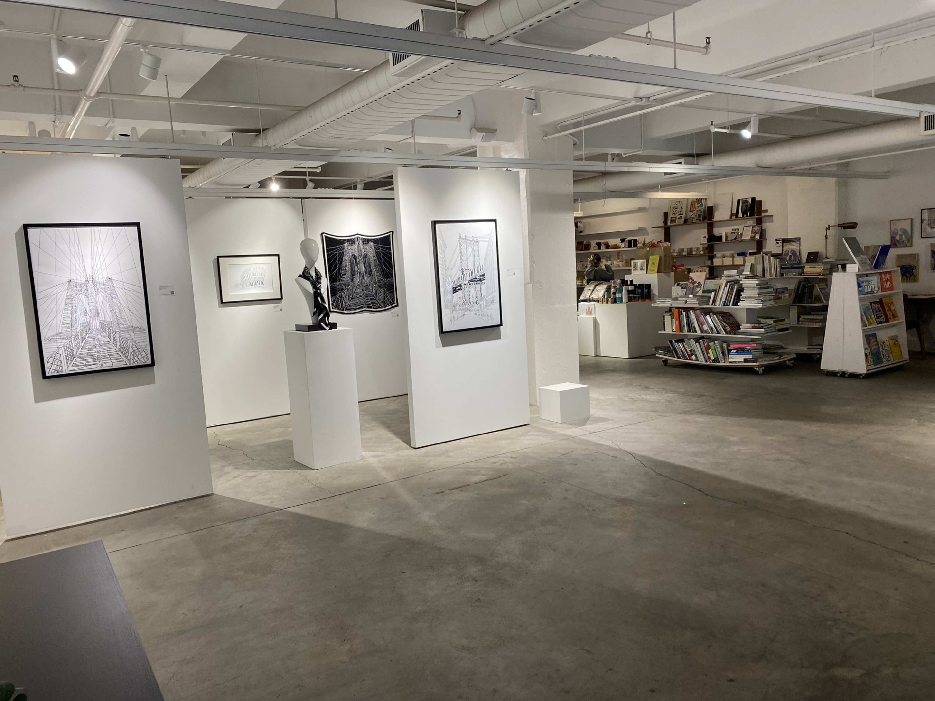 An art gallery interior with white walls displaying framed artworks and a sculpture on a pedestal, alongside a bookshelf filled with books.