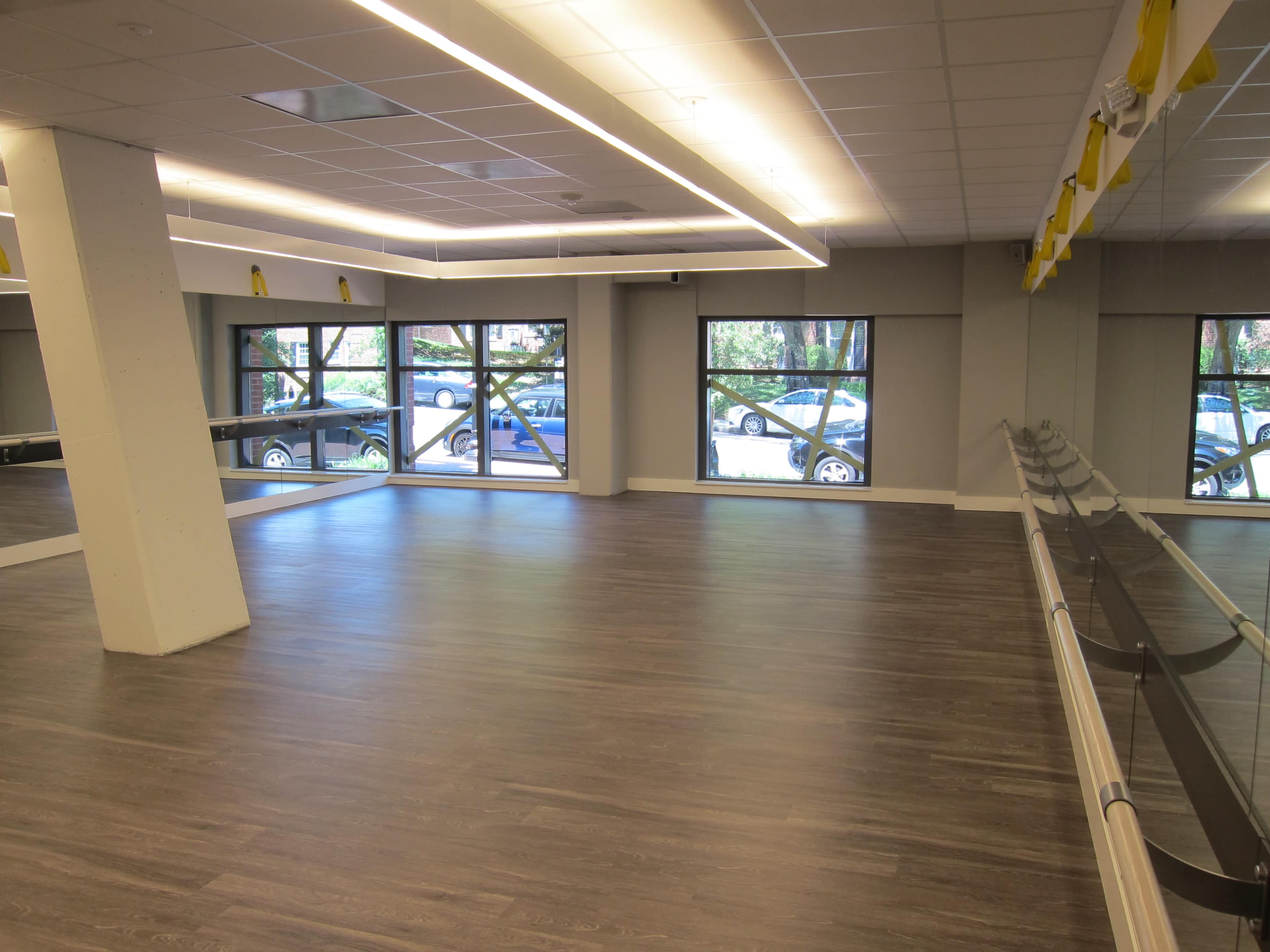 The image shows a spacious dance studio with mirrors, a barre along one wall, and large windows revealing parked cars outside.