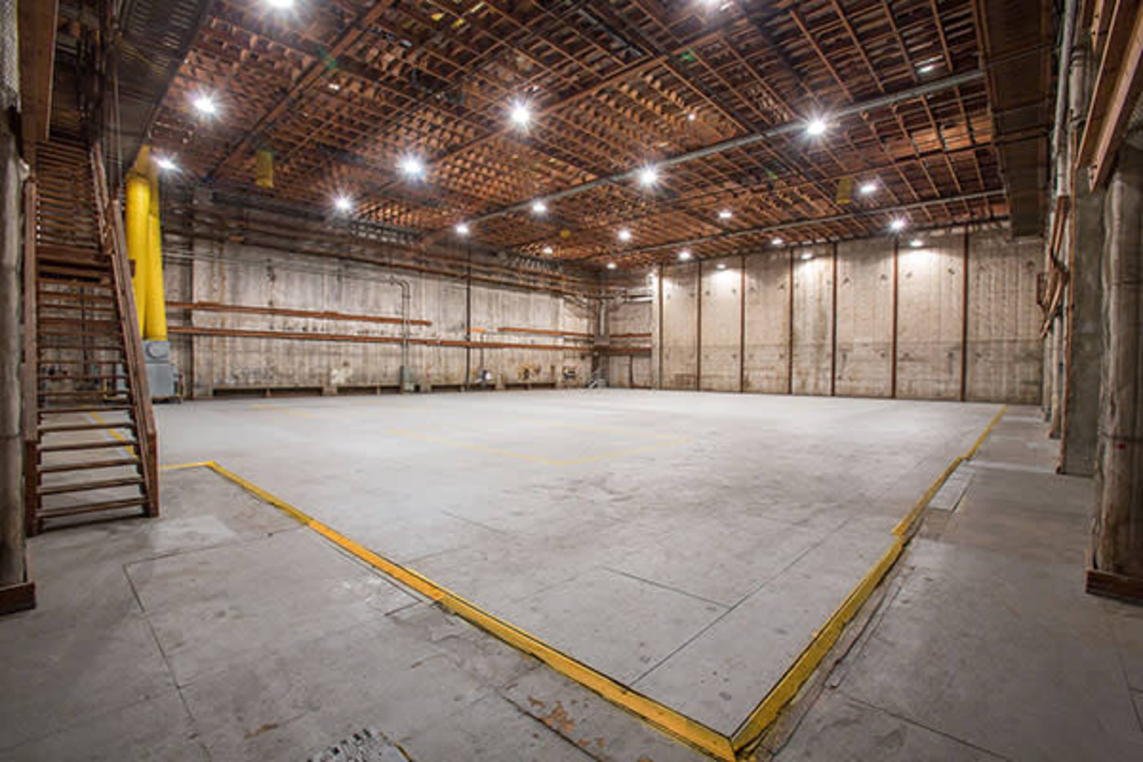 The image shows a large, empty warehouse interior with concrete floors and high ceilings supported by wooden beams.