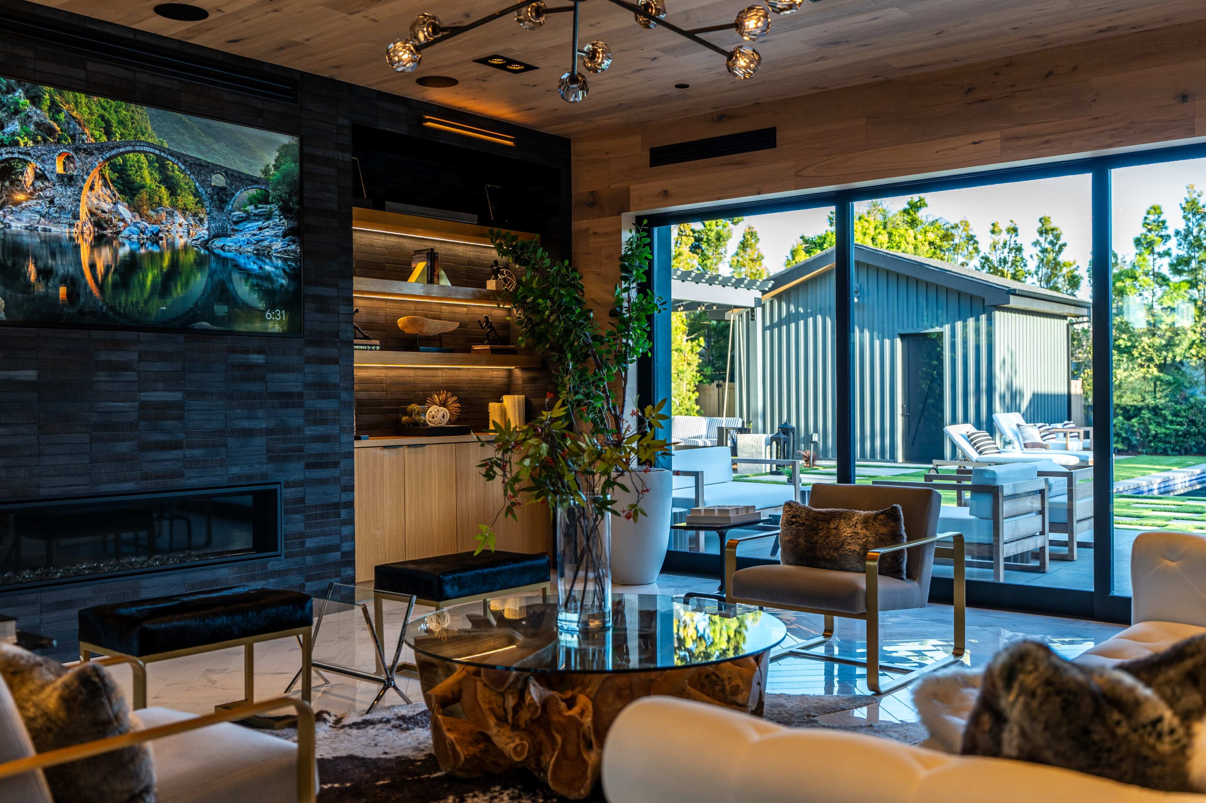 The image shows a modern living room with a large glass door opening to a patio and pool area, featuring stylish furniture and a decorative wall with a mounted screen.