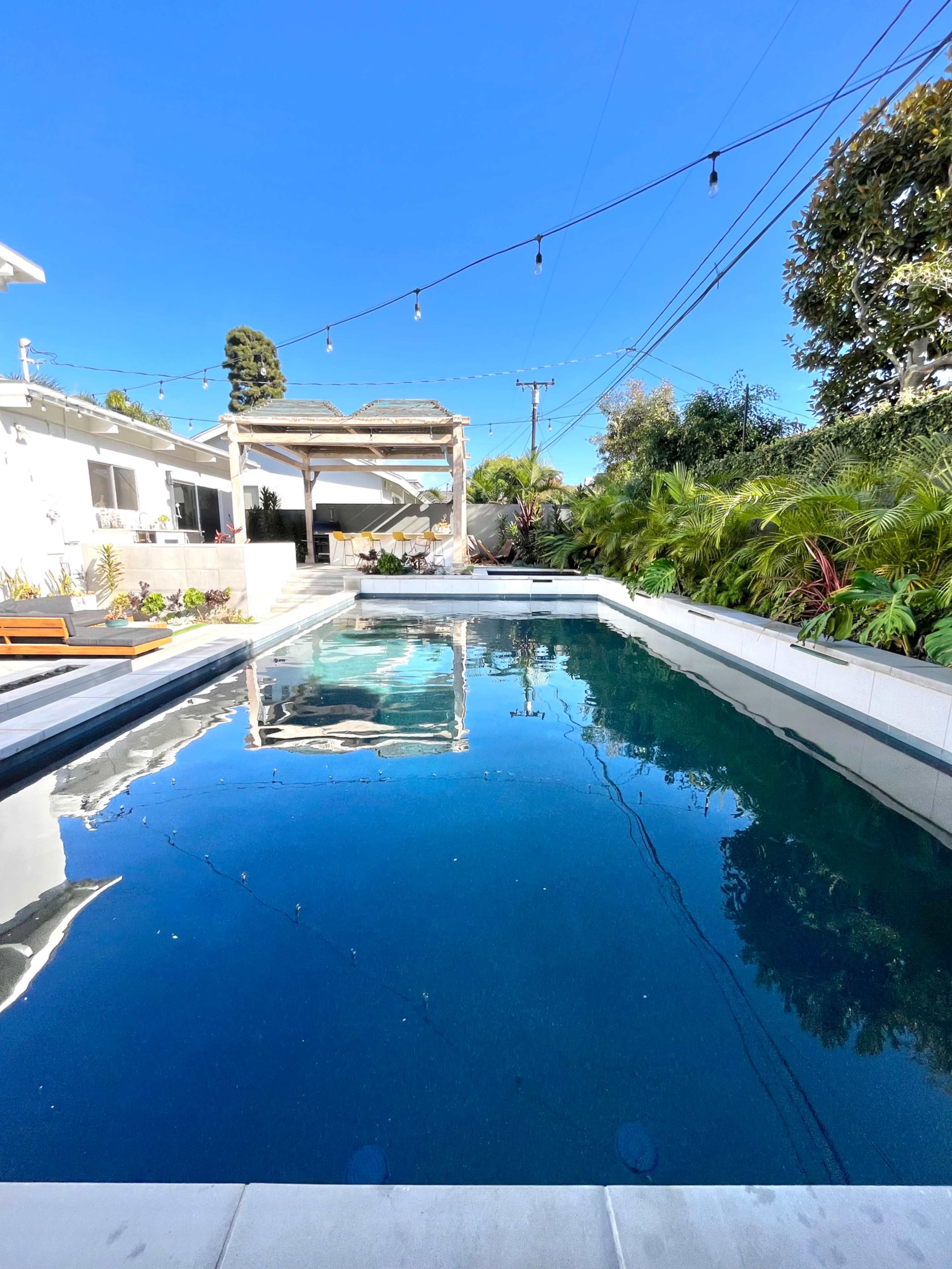 A rectangular swimming pool reflects the clear blue sky and surrounding greenery, with a pergola and string lights visible in the background.