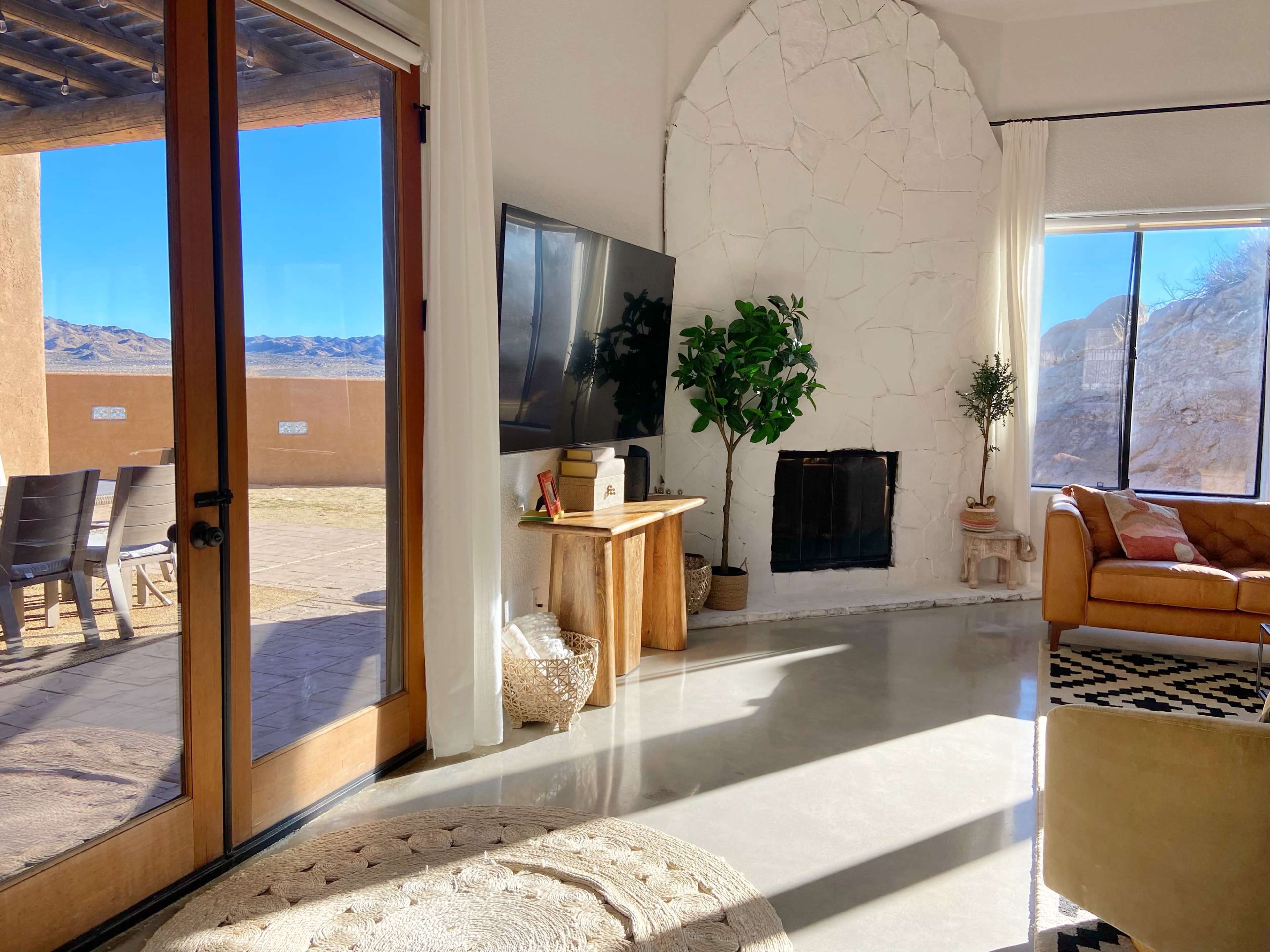 A living room features a large window overlooking a desert landscape, a stone fireplace, a television mounted on a white wall, and a tan sofa.